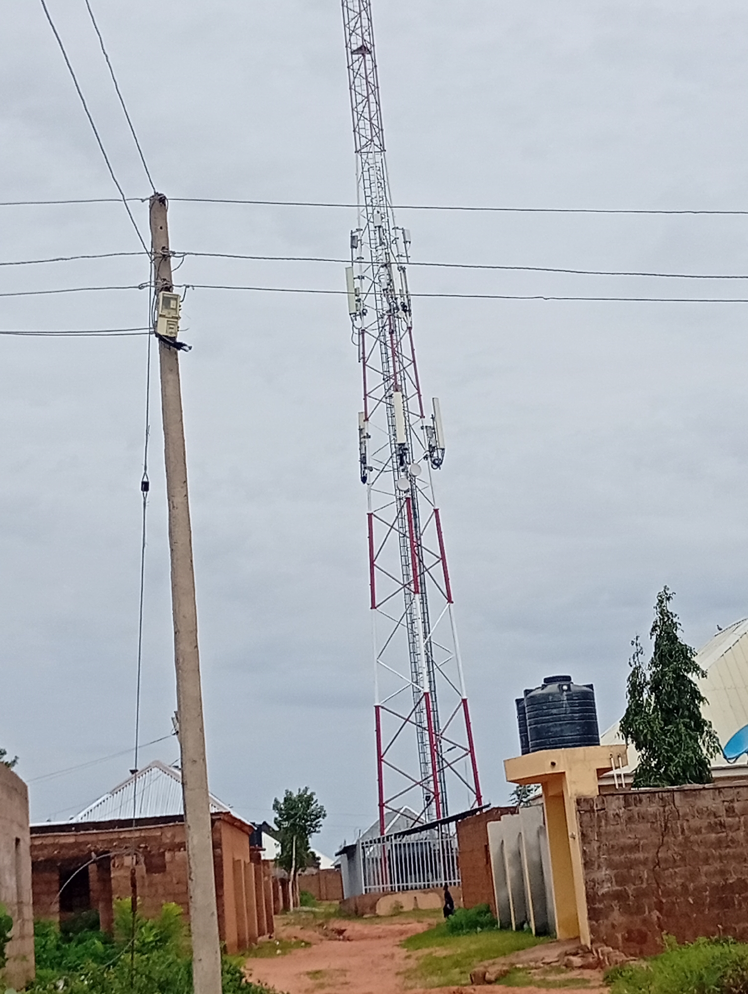 A tall, lattice-style telecommunications tower with multiple antennas is the dominant feature of this outdoor scene in Tumfure, Nigeria. The tower, painted in alternating red and white sections, rises against a cloudy sky. In the foreground, a concrete utility pole with electrical equipment attached stands to the left. Several power lines extend from this pole and across the image.
The ground is a mix of reddish-brown dirt and sparse green grass. To the right of the utility pole, a cluster of low-rise buildings with brick walls and corrugated metal roofs are visible. A large, black water tank sits atop a yellow structure, likely part of one of the buildings. A tall, conical evergreen tree is situated behind the water tank. Further to the right, a white structure with a curved roof, possibly part of a larger building or stadium, is partially visible. A few dark-colored figures, likely people, are present but too indistinct to identify clearly. One figure is walking on the dirt path in front of the buildings. The overall impression is of a semi-urban or peri-urban environment where infrastructure for communication and basic services is present.