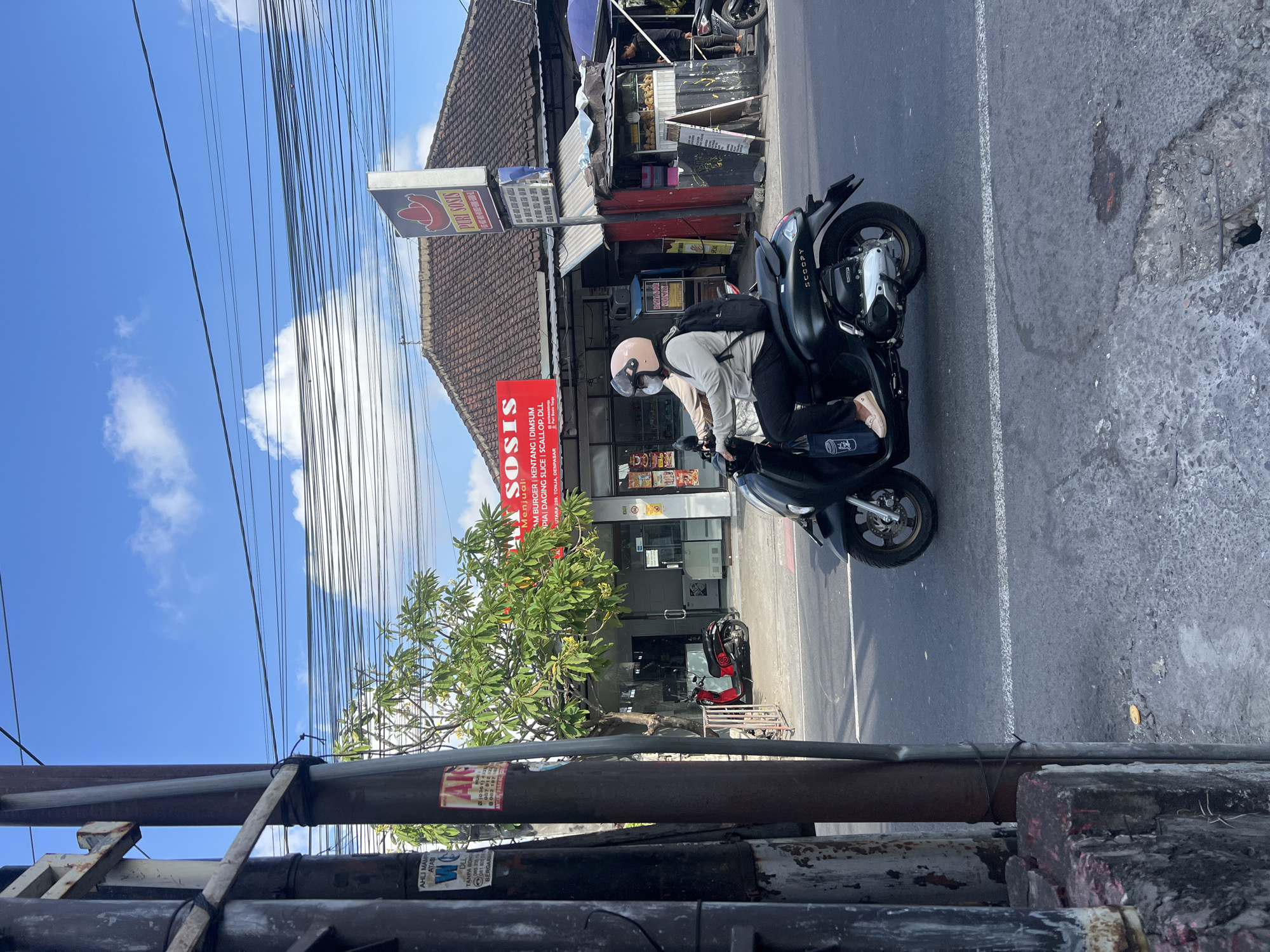 The image shows a street scene in Denpasar, Indonesia. A person is riding a black scooter on the right side of the road. The scooter has the word "SCOOPY" written on it. The rider is wearing a pink helmet and a light-colored long-sleeved shirt, dark pants, and light-colored shoes. They also have a black backpack. In the background, there is a building with a red sign that says "PURI SOSIS" and another red sign that says "SOSIS". There are several power lines overhead and a tree with green leaves. A red scooter is parked in front of the building. The sky is blue with some white clouds. The road surface appears to be asphalt and there is a white line marking the lane. There are also some concrete structures and a metal pole on the left side of the image.