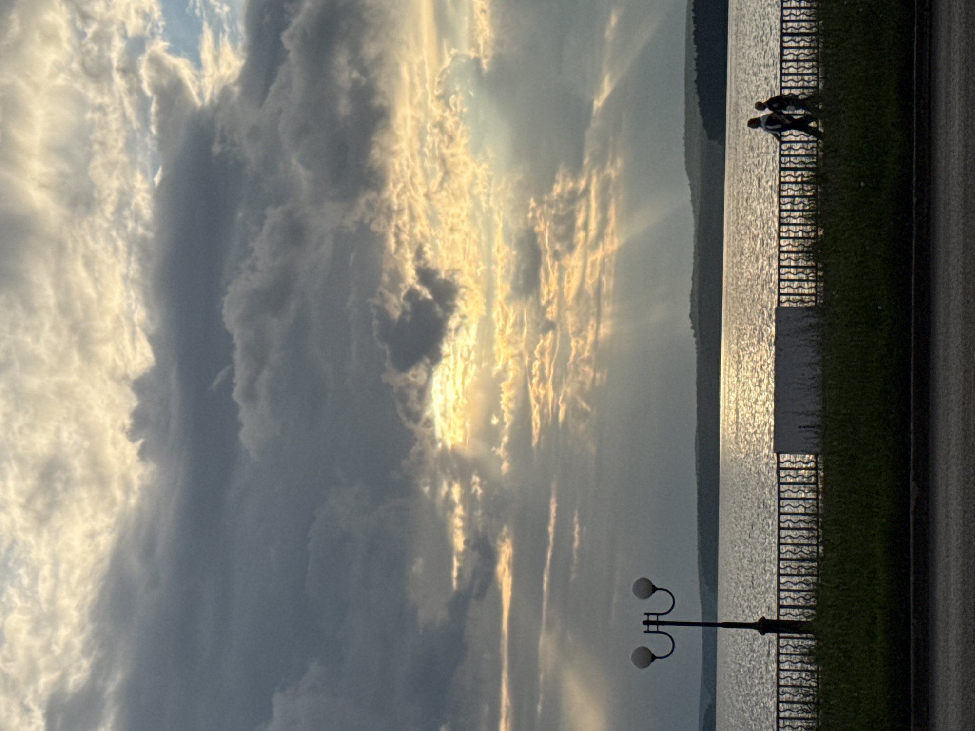 The image depicts a dramatic sky at sunset over a body of water, possibly a lake or river, in Votkinsk, Russia. The sun is setting, casting golden rays through breaks in heavy, textured clouds, creating a powerful contrast between light and shadow. The clouds are dark and voluminous on the left, transitioning to lighter, illuminated formations closer to the setting sun.
Along the waterfront, a black metal fence separates a grassy area from the water's edge. Two figures, silhouetted and appearing to be walking, are visible near the fence, adding a sense of human presence to the vast natural scene. A dual-headed street lamp stands in the foreground, its shape providing a subtle vertical element against the horizontal expanse of the water and sky.
The water is rippled and reflects the sky's dramatic light, creating a shimmering surface. The overall mood is serene yet awe-inspiring, with the natural spectacle of the sunset dominating the view. The time of day is clearly sunset, indicated by the low angle of the sun and the vibrant, warm light. The weather appears to be partly cloudy, with the clouds contributing to the striking visual effect. No text is visible in the image.