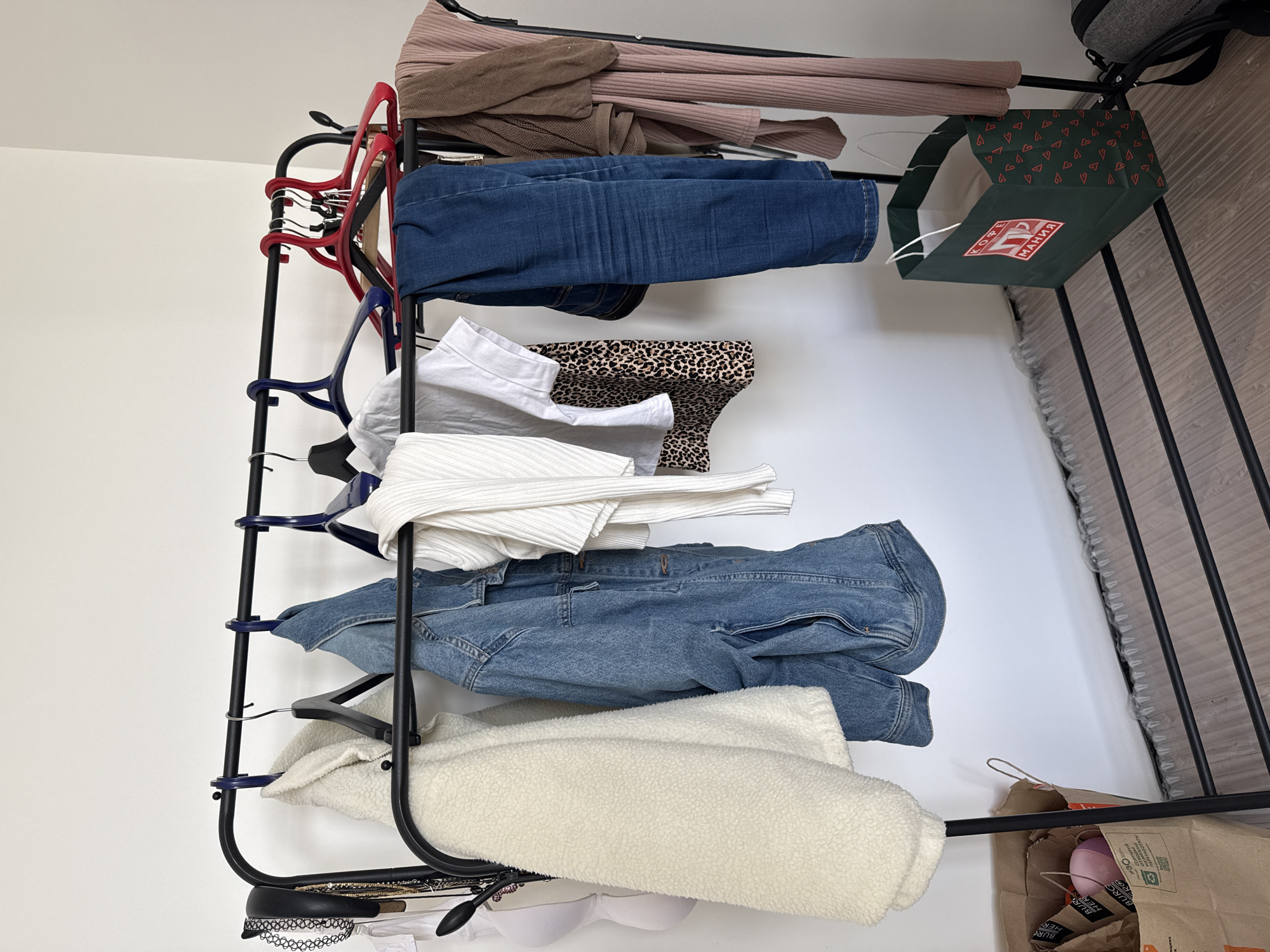 A clothes rack laden with various garments stands against a white wall. The garments, from top to bottom, include a ribbed beige knit top, a pair of blue jeans, a patterned skirt, a white blouse, a white knit top, a denim jacket, and a white fleece jacket. A green gift bag with red heart patterns and the Cyrillic word "KOPRE" is hooked on the rack. Below the rack, a small collection of accessories, including a bra and a necklace, are visible. The scene contains a bag with the word "BURGER" on the bottom right. The rack stands on a tiled floor. The location cannot be determined solely from the image.