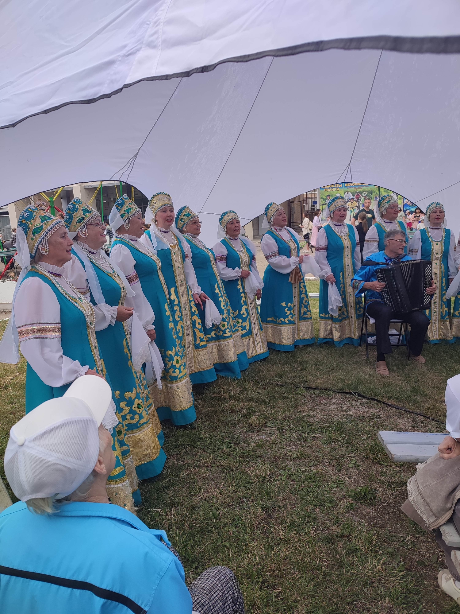 An outdoor photograph captures a group of women dressed in traditional blue and gold folk costumes, standing in a line. They are under a large white canopy. To the right, a man is seated on a chair, playing an accordion. A person in a blue shirt and white cap is in the foreground, seated and looking up at the performers. The background shows a poster with images of bicycles and text. The ground is covered in grass.