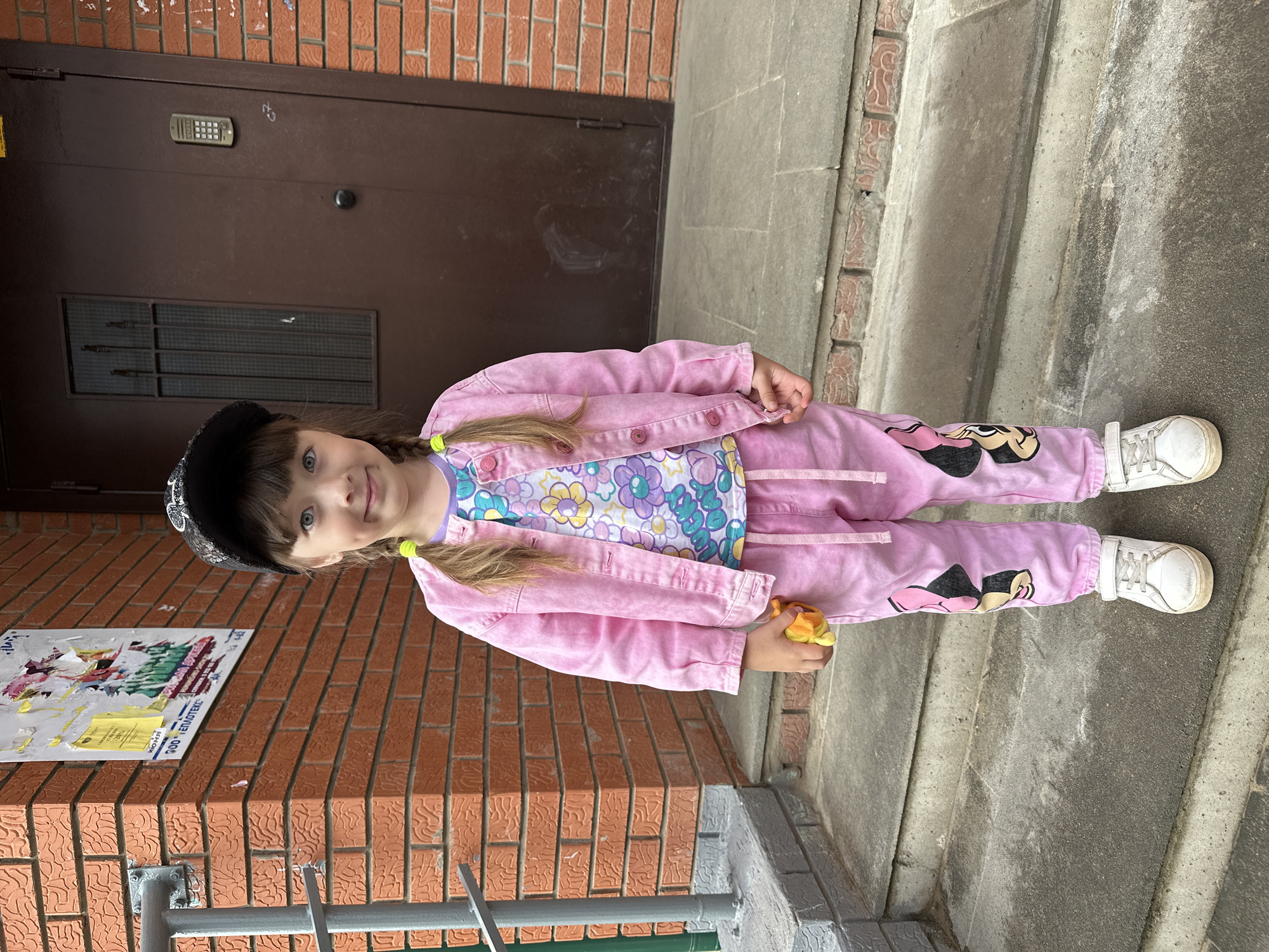 A young girl with two pigtails and a black baseball cap stands on concrete steps outside a brick building in Chaikovsky, Russia. She is wearing a pink denim jacket over a floral-patterned shirt and pink sweatpants adorned with Minnie Mouse graphics. In her right hand, she holds a small, yellow toy. The building appears to be an apartment complex, with a dark brown door and an intercom system visible on the right. A railing runs along the left side of the steps, and a bulletin board with posters is attached to the brick wall. The lighting suggests it is daytime.