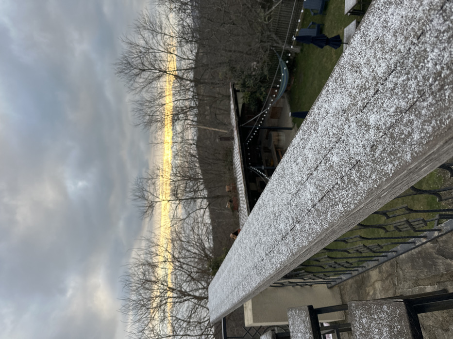 This image depicts an elevated perspective overlooking an outdoor area and natural landscape in Sighnaghi, Georgia. In the immediate foreground, a weathered wooden railing, divided into visible planks, is covered with a light, granular dusting of snow or frost. To the left, two wooden stools, similarly frosted, are partially visible. A black, ornate metal balustrade runs beneath the railing.
Below the balcony level, a green lawn or courtyard unfolds. To the left, a structure with a dark, tiled roof, possibly an open-air pavilion or kitchen, is present, featuring string lights draped across its interior space. A blue fabric hammock is strung between supports near this structure. Further right on the lawn, two dark blue outdoor chairs and two light-colored, low tables are arranged. An indistinct, reddish-brown form is partially visible behind the tiled roof structure.
The background features a dense expanse of leafless deciduous trees, characteristic of a winter or early spring season, covering a sloped hillside. A distinct, narrow horizontal band of bright yellow-orange light is visible on the distant horizon, contrasting with the heavily overcast sky dominated by various shades of grey cloud formations. No individuals are clearly discernible or engaged in specific actions.