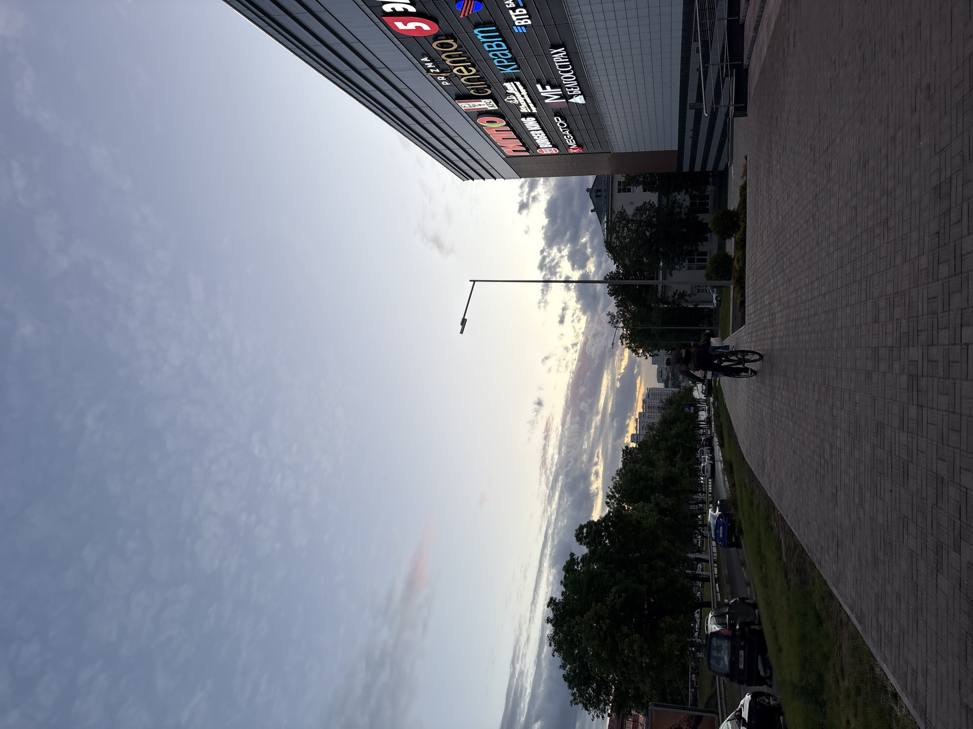 The image depicts a view in Minsk, Belarus, adjacent to a building displaying various signs, including "PRZMA CINEMA 5," "EUROPA," "KREAM," "MF," "MEGATOP," "BURGER KING," "БЕЛОСТРАХ," and "BTB". A paved walkway runs alongside the building. A person on a bicycle is visible on the walkway. Across the walkway, a row of trees borders a street with parked cars. The sky is cloudy with hints of sunset.