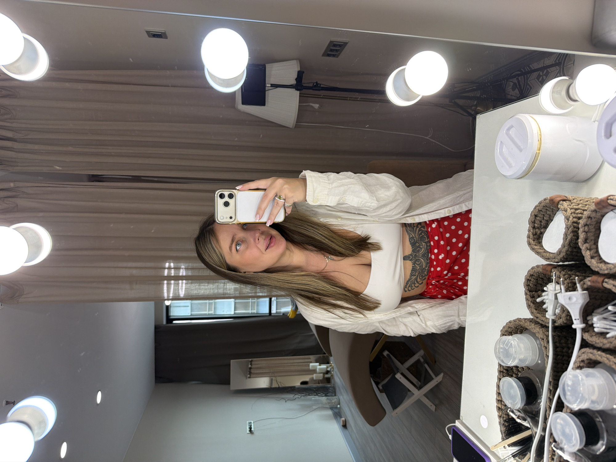 A woman is taking a selfie in a makeup room. The woman has long blonde hair, light skin, and is wearing a white crop top, a white button-up shirt, and red and white polka dot pants. She has tattoos on her left arm and chest. She is holding a white iPhone in her right hand. The makeup room has a large vanity mirror with lights around it. There are various makeup products and tools on the vanity. The background shows a glimpse of the room, including a chair, a mirror, and curtains. There is no evidence in the image to determine if the location is in Moscow, Russia.
