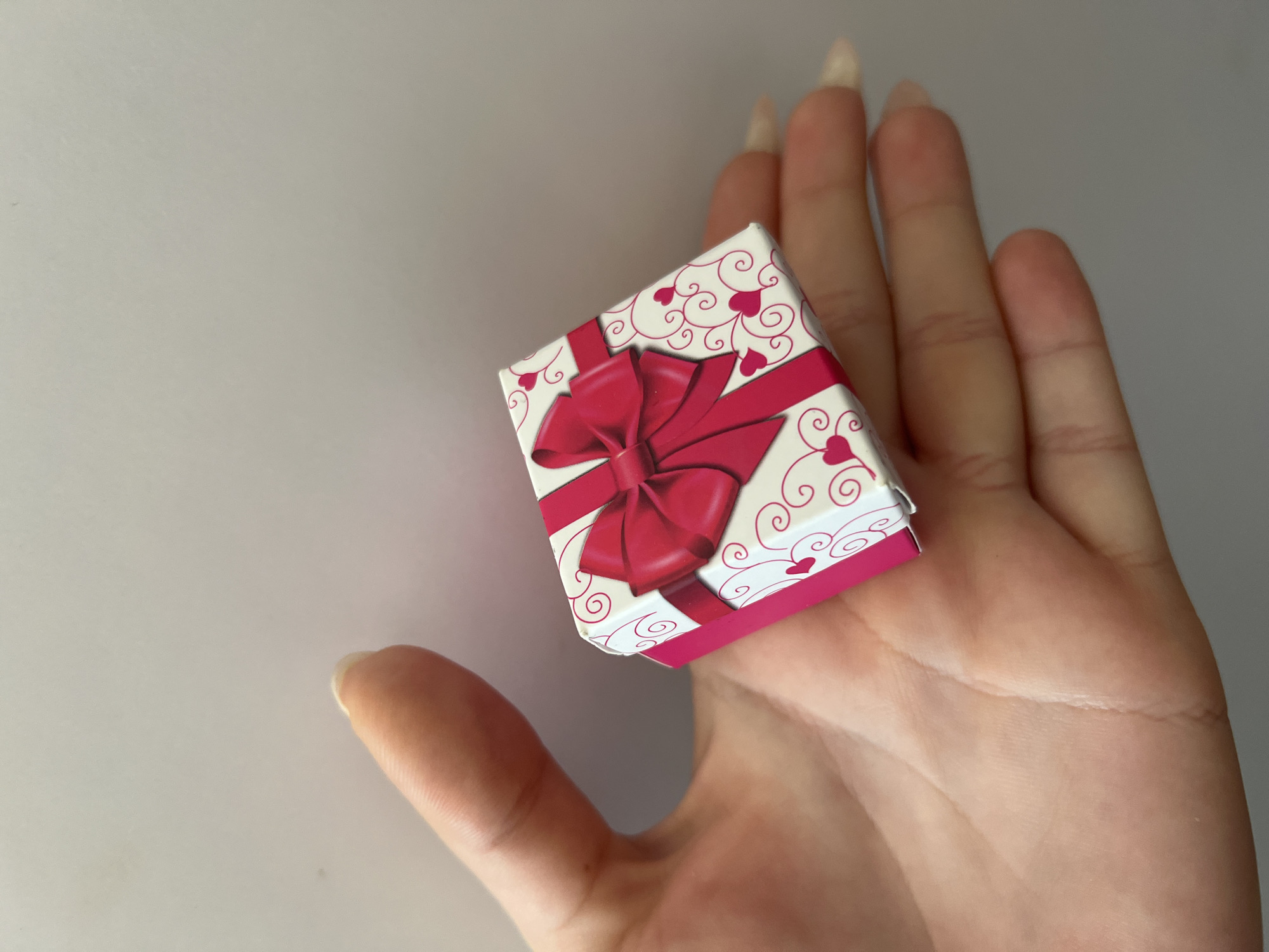 A human hand, with neatly manicured nails, cradles a small, square gift box. The box is white with a pink ribbon and a pink bow on top, and features a pattern of swirling lines and small pink hearts. The background is a plain, light gray. The image appears to be a close-up shot, focusing on the box and hand.
