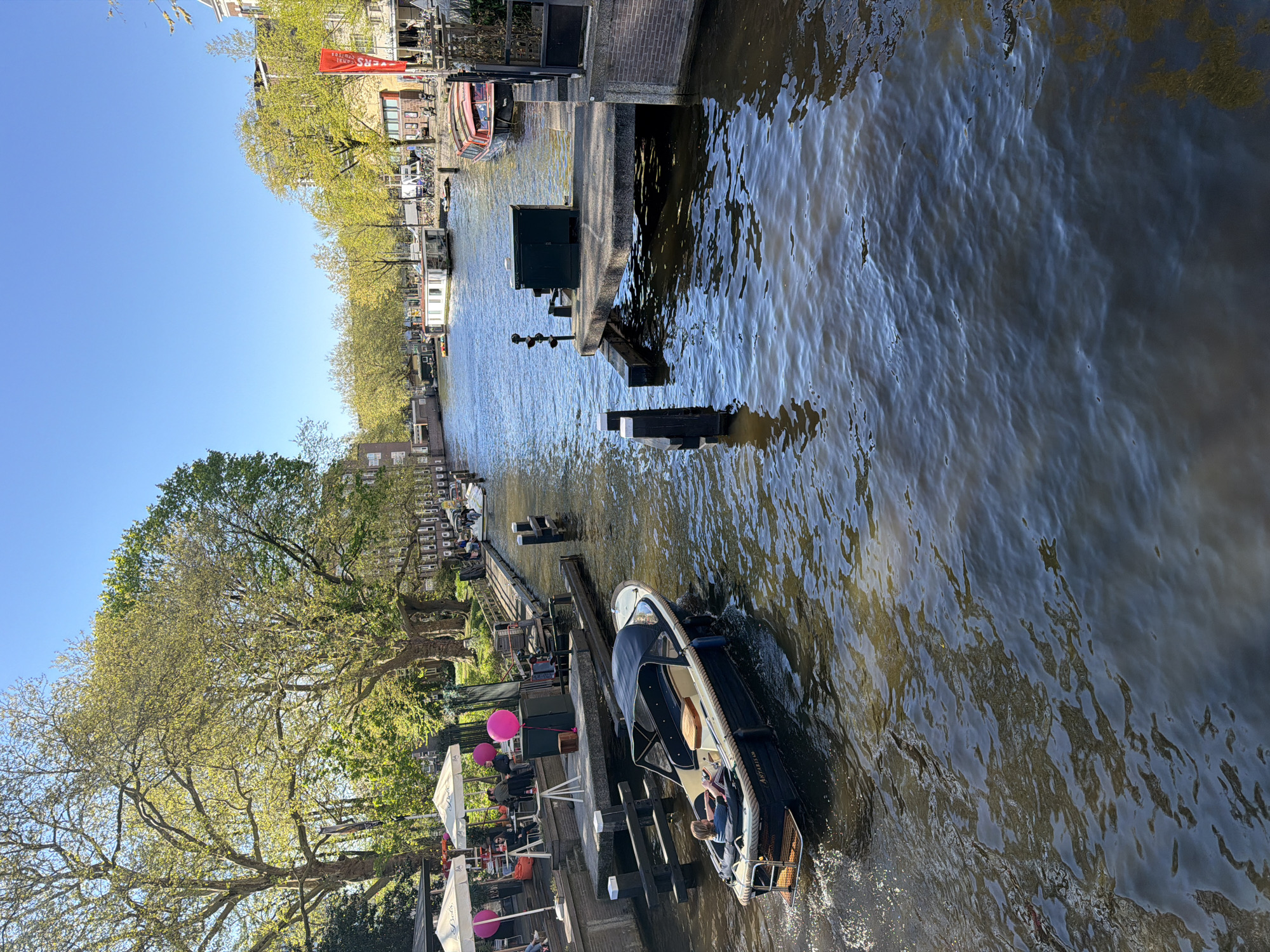 This image captures a bright, sunny daytime scene along a canal in Amsterdam, characterized by leisurely activity and urban charm.
In the foreground, a dark blue motorboat with a light-colored hull and a canopy travels from right to left, generating a small wake. On its side, the name "Amsterdam" is clearly visible, and at least two people are onboard, one seemingly steering while another sits near the bow. The canal water is dark and choppy, reflecting the clear blue sky above.
The left bank features a vibrant promenade shaded by mature trees with fresh, green spring foliage. An outdoor cafe or restaurant is set up here, with several people seated at tables under white and prominent pink umbrellas, enjoying the pleasant weather. Further down this bank, a white houseboat is moored.
The right bank presents a more industrial edge with a brick wall, sturdy wooden mooring posts, and a dark green utility box at the water's edge. Beyond these structures, additional boats, including a distinct red one, are moored along the canal.
Traditional multi-storied buildings, characteristic of Dutch architecture, are visible behind the trees and along both canal banks. The sky overhead is a brilliant, cloudless blue. A red banner, partially visible on the far left bank, explicitly reads "CANAL CRUISES," serving as a clear location cue.
The scene depicts a calm yet active atmosphere, with a boat tour or private leisure craft cruising the waterways while patrons relax at a waterside establishment, likely during the mid-morning or afternoon due to the strong sunlight and minimal shadows.