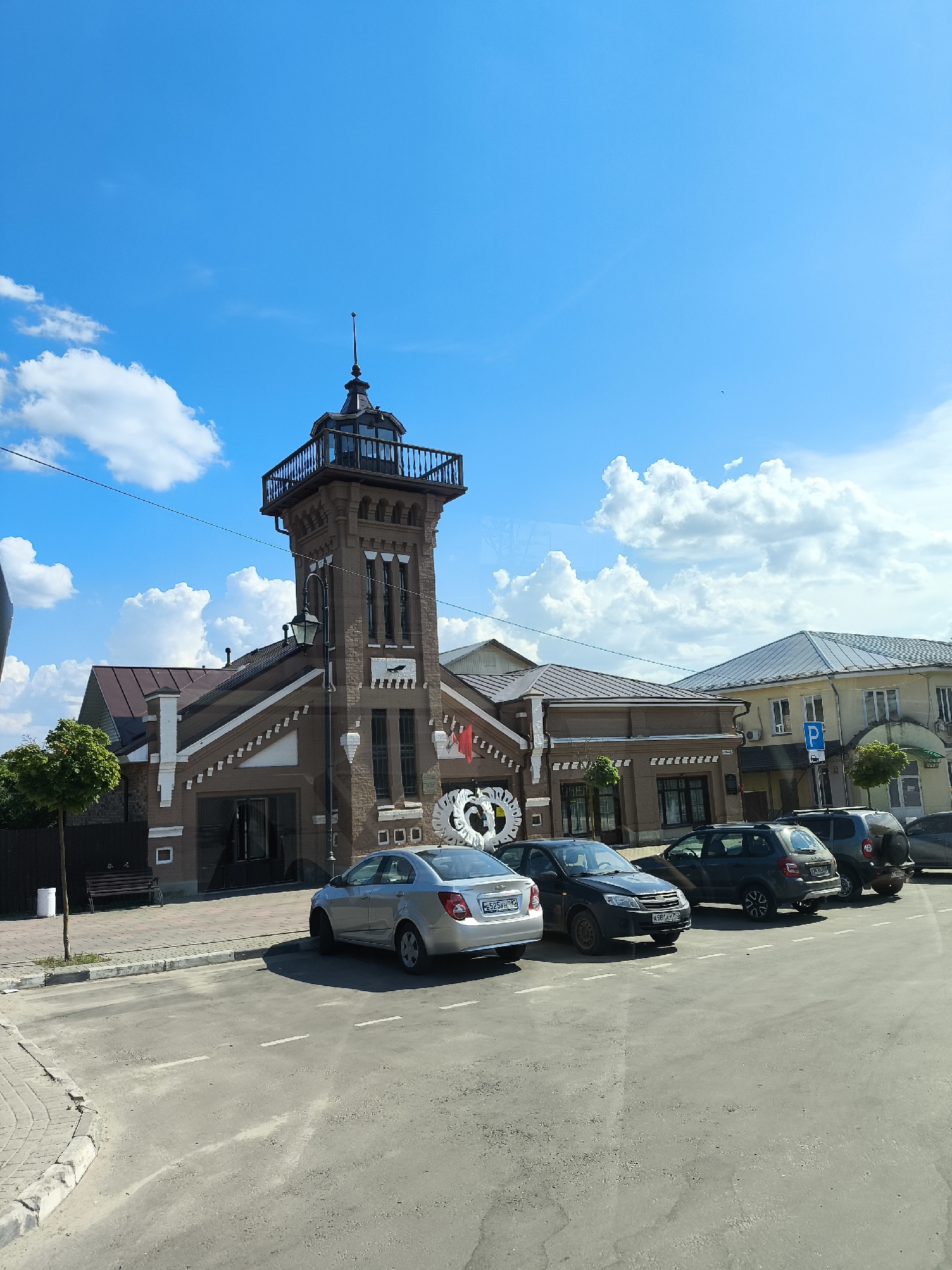 The image shows the historic fire tower building in Taldom, Russia, on a clear day with a bright blue sky and scattered white clouds. The fire tower is a prominent, multi-story brick structure with a dark roof and a lookout platform at the top. In front of the building, a paved area serves as a parking lot for several cars, including a silver sedan, a dark hatchback, a dark SUV, and another dark SUV. The fire tower has several windows, decorative white brick accents, and a red flag displayed on its side. Adjacent to the fire tower are other buildings with similar architectural styles, featuring tiled roofs. A small, manicured tree stands to the left of the fire tower, and a wooden bench and a trash can are visible on the sidewalk. The surrounding environment appears to be a well-maintained urban area.