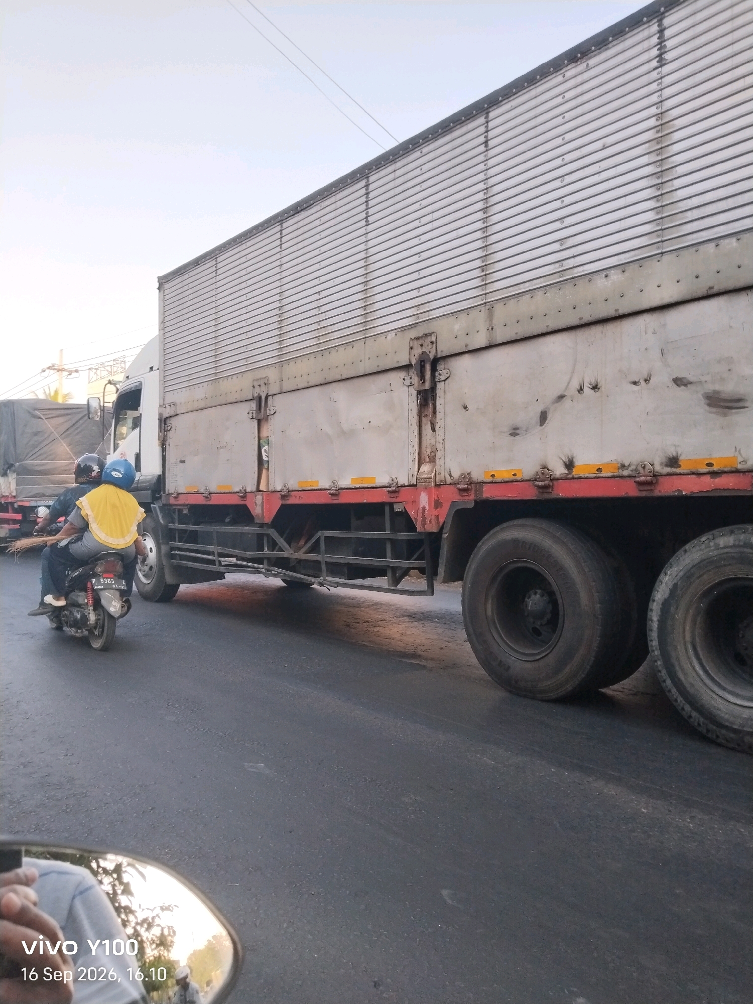 Two individuals on a motorcycle are captured in motion on a paved road, passing by the side of a large, silver semi-trailer truck. The truck's cargo container features a ribbed metal exterior with visible rivets and some minor staining or scuff marks. It also has red trim along its lower edge, punctuated by yellow reflective markings. The rear wheels of the truck are prominent, showing dual tires on the right side of the frame. The sky above is a pale blue. In the lower right corner of the image, a reflection in what appears to be a motorcycle's side mirror shows a person's hands and a portion of a blue shirt, along with some greenery and sky. The timestamp on the reflection reads "vivo Y100 16 Sep 2026, 16.10". The presence of the timestamp and the context suggest this photograph was taken in Situbondo, Indonesia.