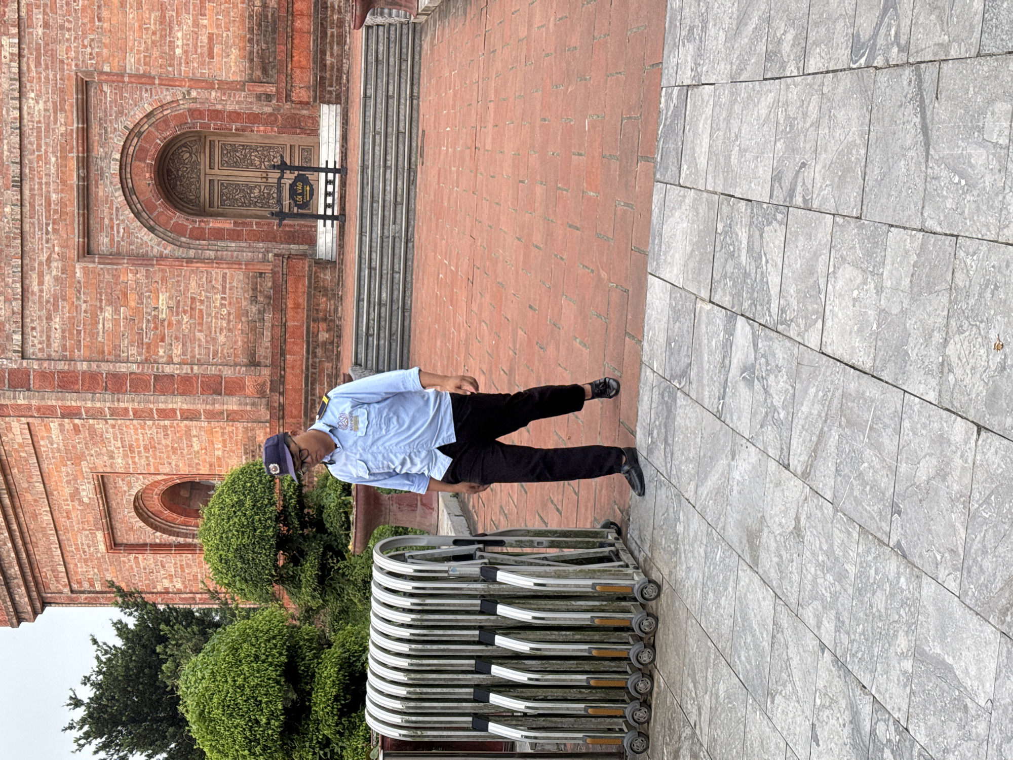 A security guard, wearing a light blue collared shirt, dark pants, and a dark cap, walks down a brick pathway in front of a large red brick building with an arched wooden door. The guard is carrying a badge on their shirt. To the left of the guard, a silver retractable gate stands closed, its sections upright and resting on small wheels. The pathway is bordered by manicured green bushes on the left and a grey stone path on the bottom. A sign with Vietnamese text and an arrow points towards the building's entrance. The building has architectural details around the doorway and along its facade. The setting appears to be an outdoor public space in Tây Hoa Lư Ward, Vietnam, likely a historical or cultural site given the architecture.