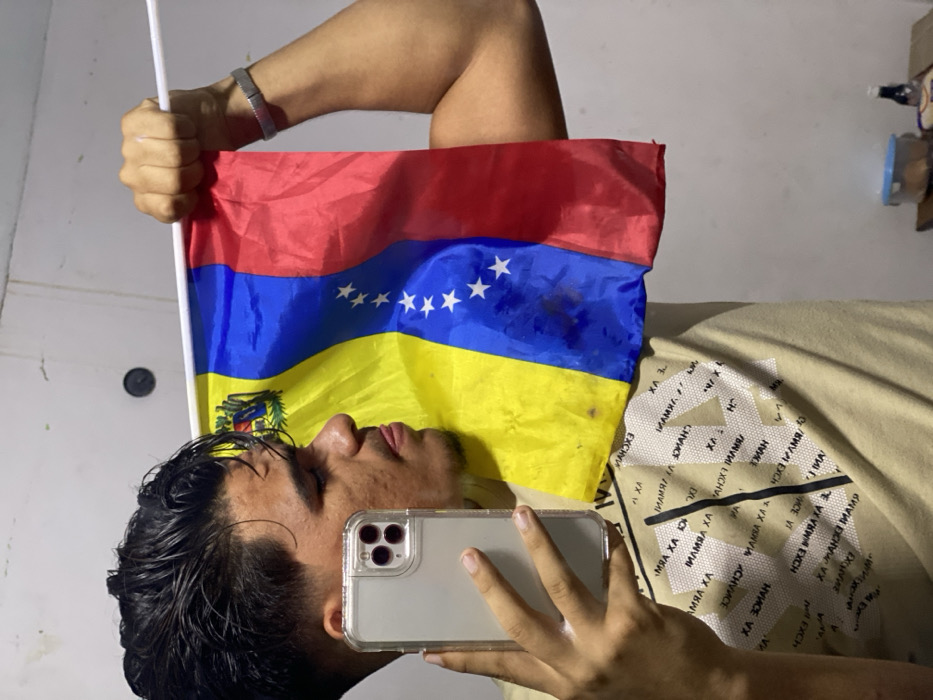 A person lies down, holding a smartphone and a Venezuelan flag. The flag is red, blue, and yellow with seven stars in the blue stripe. The person is wearing a light brown t-shirt with a repetitive pattern of "AX EXCHANGE". The phone is in a clear case, and the person's wet hair is visible. A white wall and some objects are visible in the background.