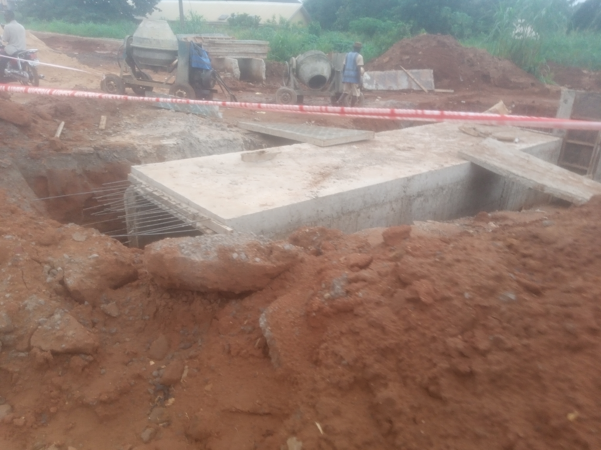This is an outdoor, medium-shot, eye-level photograph of a construction site in Nawfija, Nigeria. The image depicts a partially excavated area filled with red soil and concrete structures. In the foreground, mounds of red soil dominate the scene. A large, rectangular concrete slab sits in the middle ground, partially covering an open pit that is reinforced with rebar. To the left of this slab, more red soil is visible, along with the edge of a deeper excavation. In the background, two concrete mixers are present. One is a larger, stationary unit, while the other appears to be a mobile cement mixer on wheels. A person in a blue vest and beige shorts is standing near the mobile mixer, and another individual on a motorcycle is visible to the far left of the image. A red and white caution tape stretches across the scene, indicating a safety perimeter. The environment appears to be a dusty and unfinished construction area, with some greenery visible in the distance.