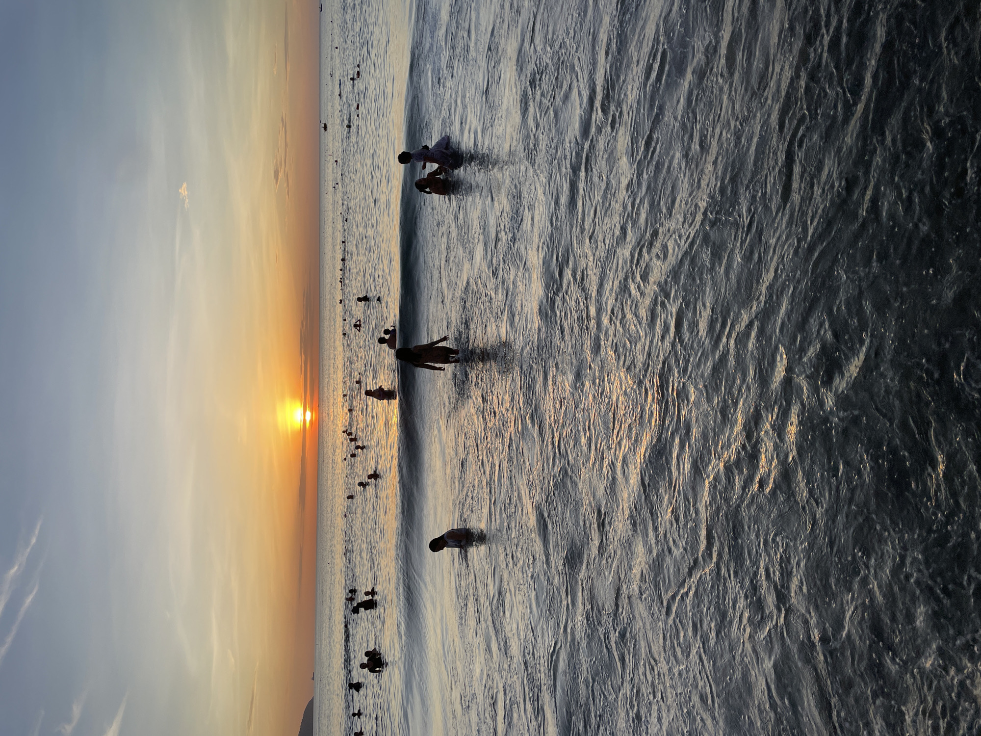 The image captures a sunset scene at a beach in Da Nang, Vietnam, likely My Khe Beach due to its popularity for sunset viewing and its expansive shoreline. The sun is low on the horizon, casting a warm orange glow across the sky and water, with a few wispy clouds present. Numerous people, including adults and children, are visible in the shallow water, some wading and others swimming. The waves are gentle, creating ripples and reflections on the surface. In the distance, a faint outline of land or mountains is visible, characteristic of the Da Nang coastline. The overall atmosphere is one of leisure and enjoyment of the natural beauty of the beach at dusk.