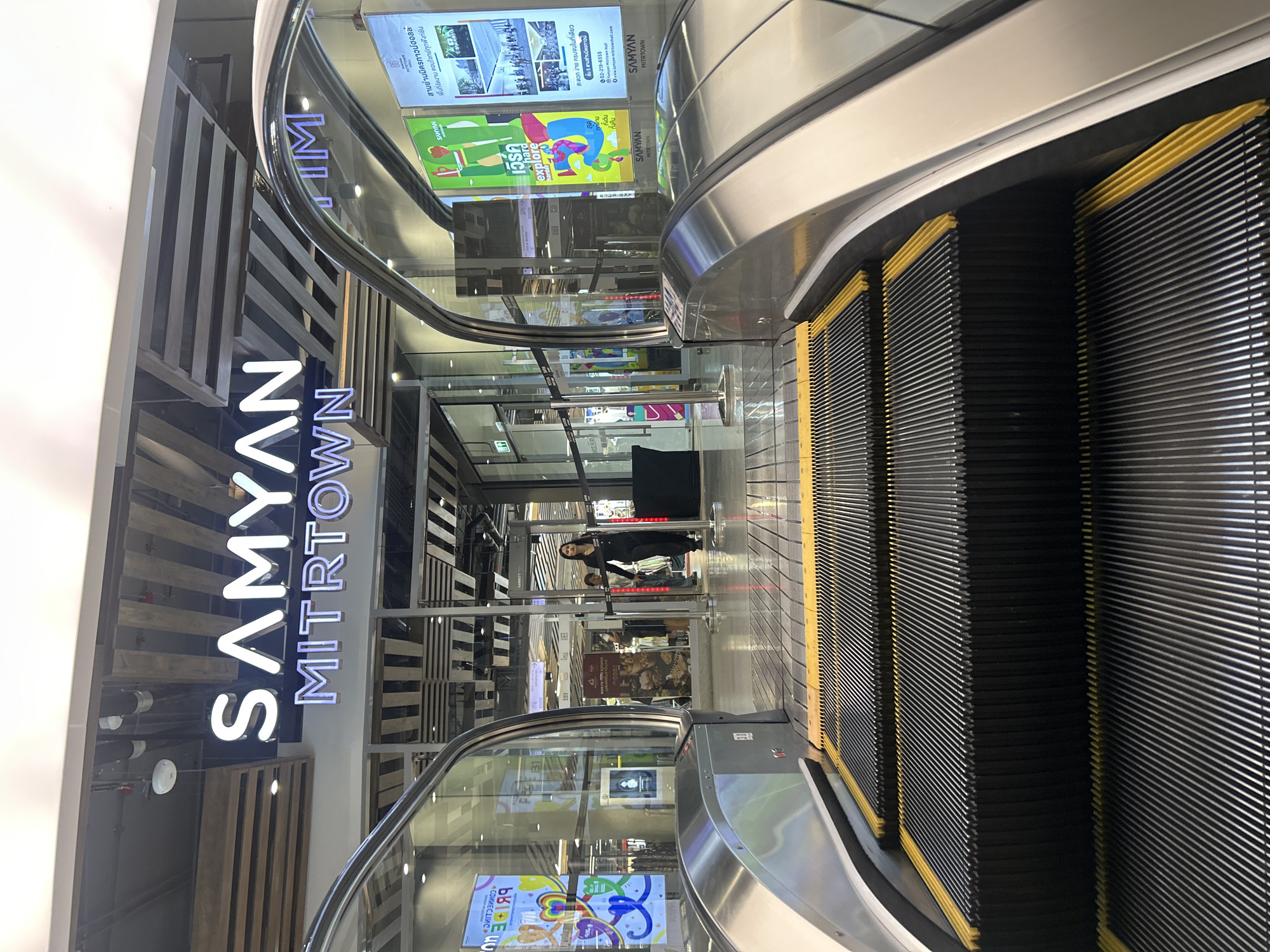 The image shows the entrance of Samyan Mitrtown, a shopping mall in Bangkok, Thailand. In the foreground, the steps of an escalator are visible, with yellow safety lines. To the left and right, the curved glass enclosures of escalators are present. Behind the escalators, the illuminated sign for "SAMYAN MITRTOWN" is prominently displayed. A woman in black clothing is walking through a glass-doored entrance in the middle ground. Several posters and advertisements are visible on the walls and glass panels of the mall, some in Thai script. The environment appears to be an indoor, well-lit public space, likely a mall.