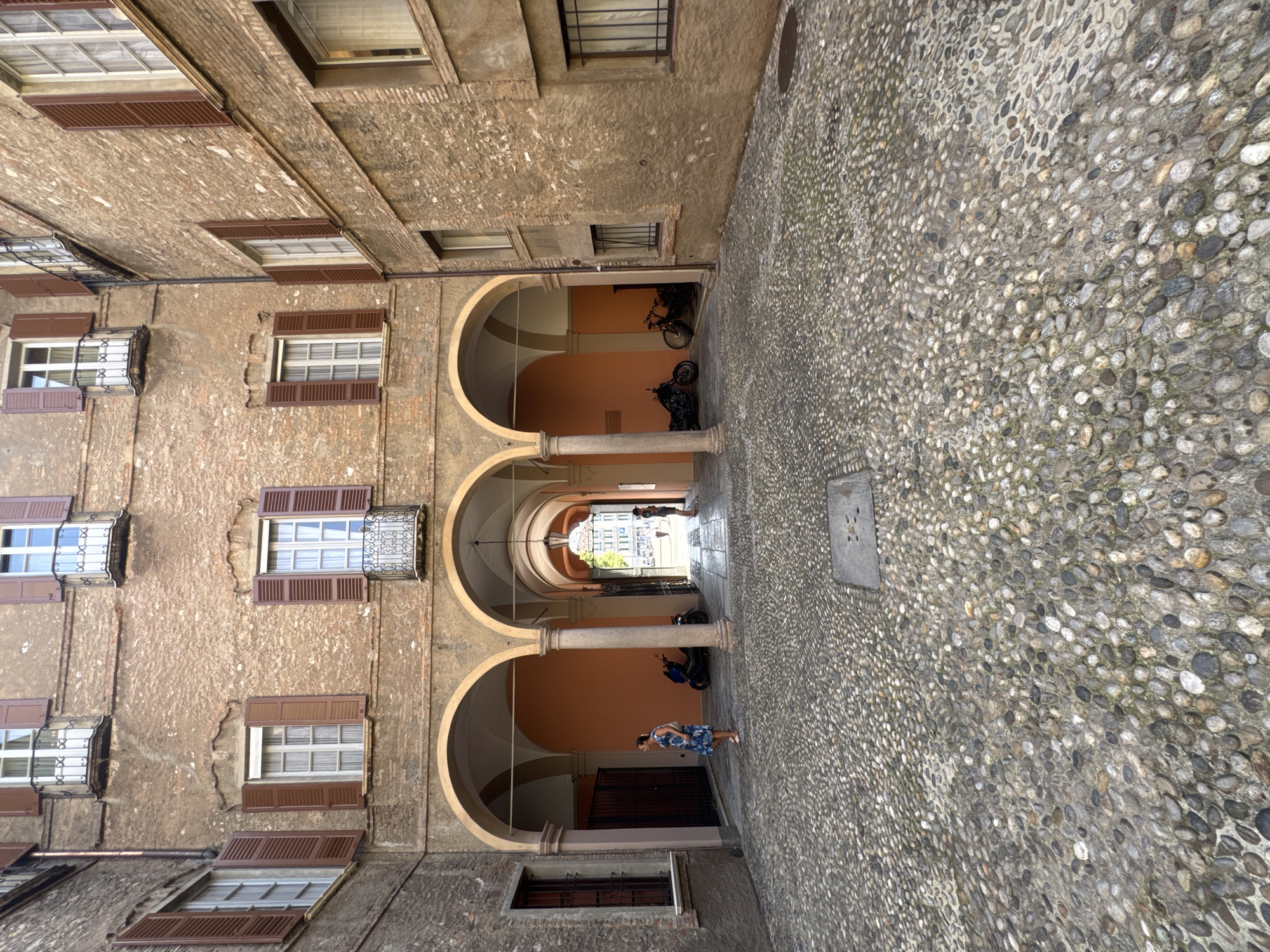 This is a photograph of a cobblestone courtyard surrounded by buildings with an arched walkway leading to another area. The buildings are constructed of brick with stucco and feature numerous windows with brown shutters. In the foreground, a woman in a blue and white floral dress is standing on the cobblestones, facing away from the camera. To her left, there is a dark metal gate. Further into the courtyard, under the arched walkway, a blue scooter is parked. Beyond the scooter, a black motorcycle and a bicycle are visible. In the distance, through the arches, a street scene is visible with more buildings and people walking. The lighting suggests it is daytime, possibly late afternoon, with shadows indicating the sun's position. The overall atmosphere is one of an old European city. This location is identified as the Piazza della Riforma in Lugano, Switzerland.