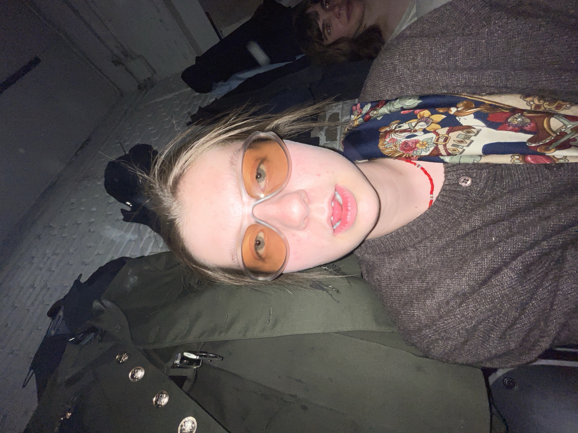 The image shows a selfie-style shot of a person with light skin, wearing orange-tinted glasses and a brown sweater with a colorful scarf draped around their neck. A red beaded necklace is visible on the neck. The person's mouth is slightly open. In the background, partially visible, is another person with dark hair. The setting appears to be an indoor space with a stone-like floor and wall, possibly a basement or similar rough, enclosed environment. The person is leaning on a green object that may be a military-style coat or bag, which has metal buttons. It is not possible to determine the location in the city of Moscow from this image.