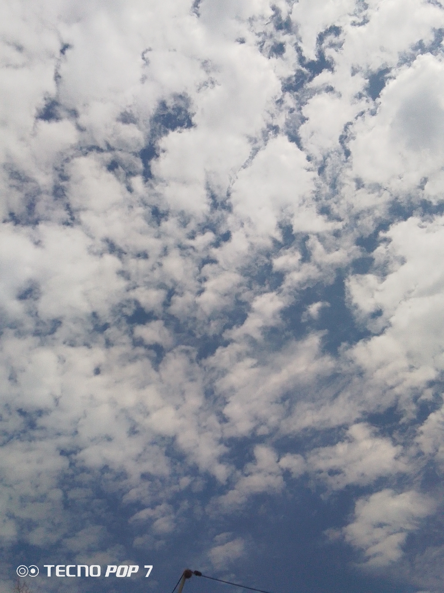 The image primarily features a daytime sky filled with scattered white and greyish clouds against a vibrant blue background. The clouds vary in shape and density, ranging from smaller, wispy formations to larger, more defined cumulus-like structures, with ample patches of blue sky visible between them. This suggests a partly cloudy, fair weather condition. At the very bottom edge of the frame, a small part of a dark utility pole or street light fixture is visible, along with some thin wires, indicating an outdoor setting. No people are present, and no specific activities are taking place. The time of day appears to be bright, likely mid-day. In the lower left corner, the text "OO TECNO POP 7" is clearly visible, likely a camera watermark. The location is Altagracia, Venezuela.