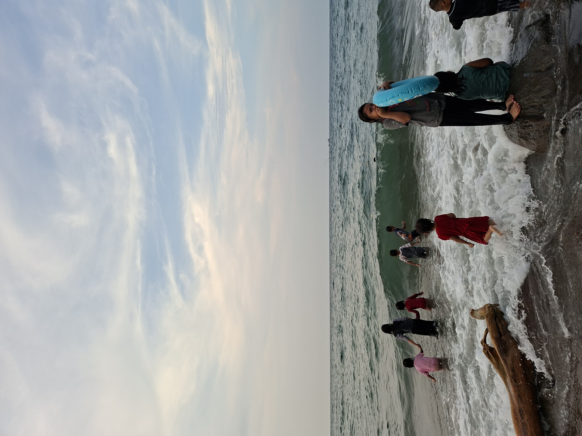 The image depicts a group of people, primarily children, enjoying the sea at a beach in Lhokseumawe, Indonesia. On the right half of the frame, the ocean's edge is visible with foamy waves washing onto the shore. Several children are playing in the water, some fully immersed while others are standing closer to the beach. The children are dressed in casual clothes, including dresses, shirts, and pants, in various colors like red, pink, and grey. One person is holding a blue inflatable ring. A large piece of driftwood is partially submerged near the shoreline. The left half of the image showcases a predominantly clear sky with wispy clouds. The sky transitions from a pale blue to a lighter tone towards the horizon. The overall setting suggests a recreational scene at a beach.