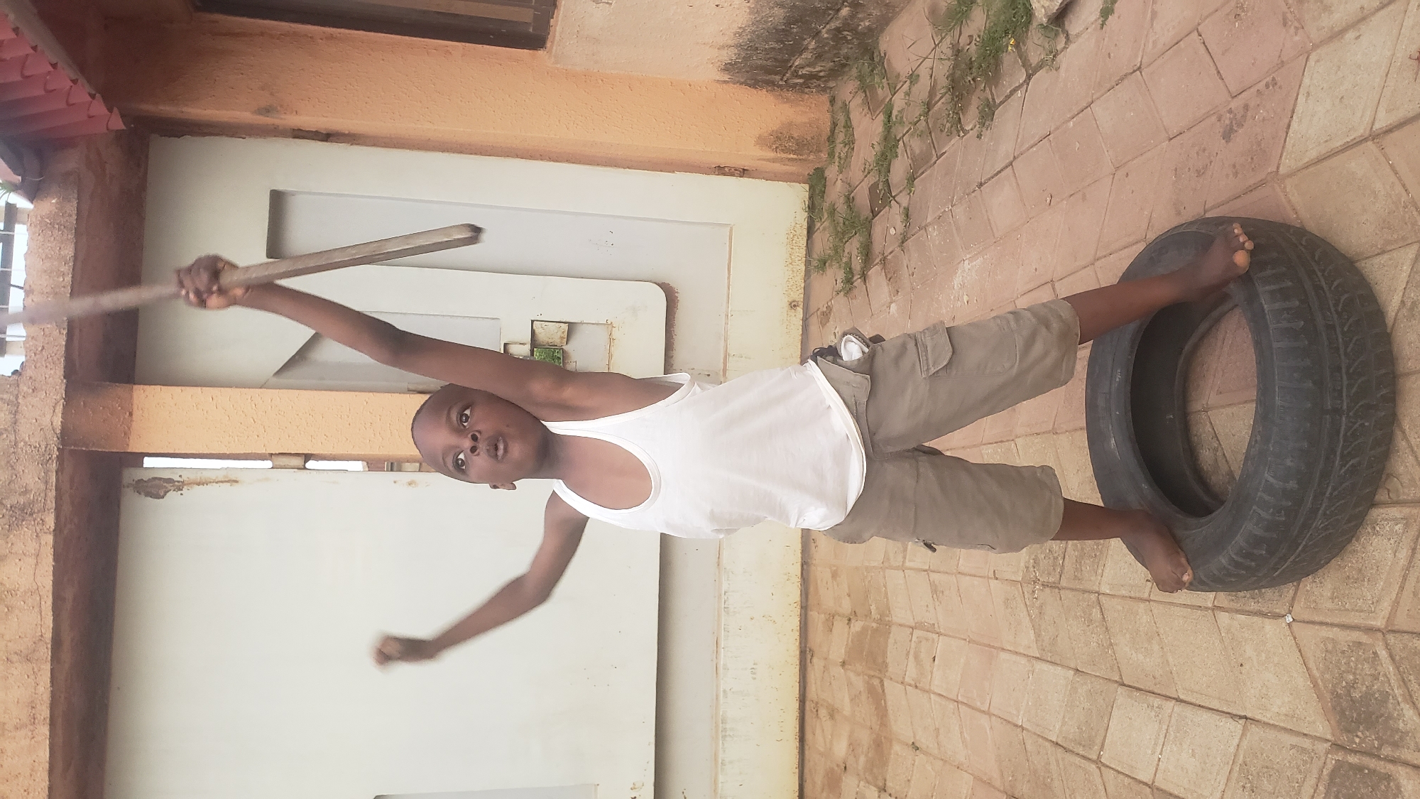 A young boy stands with his feet on a tire in Sabon Tasha, Nigeria. He is wearing a white tank top and khaki cargo shorts. He is holding a wooden stick above his head with both hands, with his arms extended. His mouth is open, and he appears to be yelling or singing. The background consists of a concrete wall and a metal gate. The ground is paved with square tiles. The lighting suggests it is daytime.