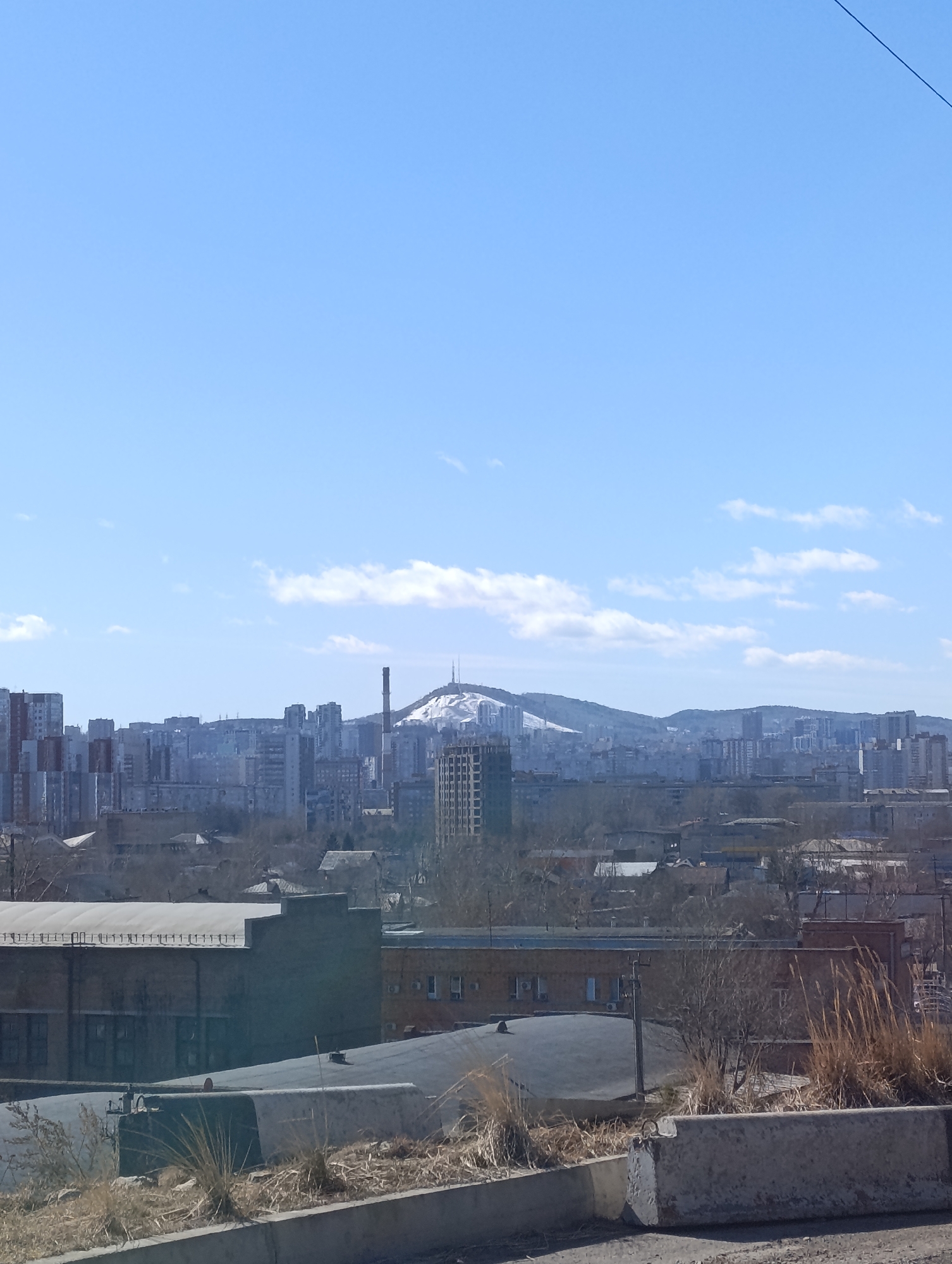 The image presents a daytime cityscape of Krasnoyarsk, Russia. The foreground features concrete barriers and dry vegetation. Mid-ground shows industrial buildings and low-rise structures interspersed with trees. In the background, a skyline of high-rise buildings is visible, with a mountain ridge partially covered in snow. A tall smokestack stands out among the buildings. The sky is blue with scattered clouds.