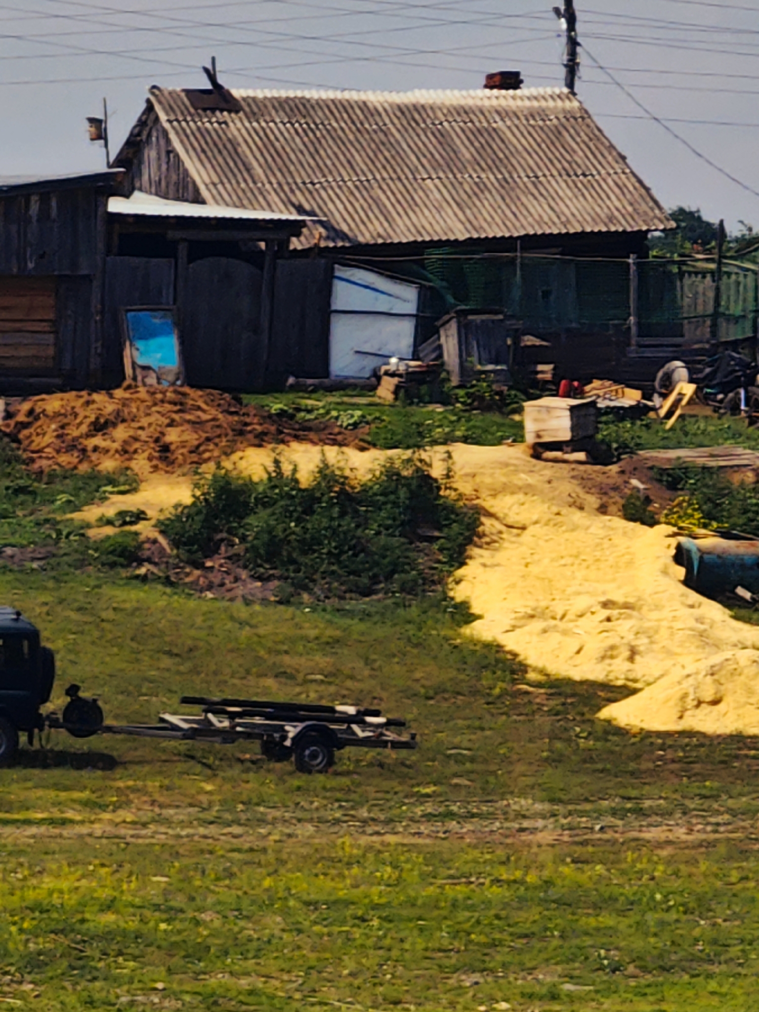 The image captures a rural scene in the village of Pogodaevo, Russia. In the background, a weathered, wooden building with a corrugated metal roof stands prominently. To its left, a smaller wooden structure is attached. In front of these buildings, a large pile of organic material, possibly straw or hay, is visible, along with a bright blue object that appears to be a discarded piece of plastic or metal. A portion of a yellow mound, possibly sand or a similar material, extends into the mid-ground.
In the foreground, a dark-colored vehicle, likely a truck, is partially visible on the left side. Attached to this vehicle is a trailer carrying several long, dark objects, possibly pipes or lumber. The ground is covered in green grass, with some patches of dirt and dry vegetation. The sky above is overcast. The overall impression is one of a utilitarian, perhaps agricultural or construction-related, outdoor space. There are no people visible in the image.