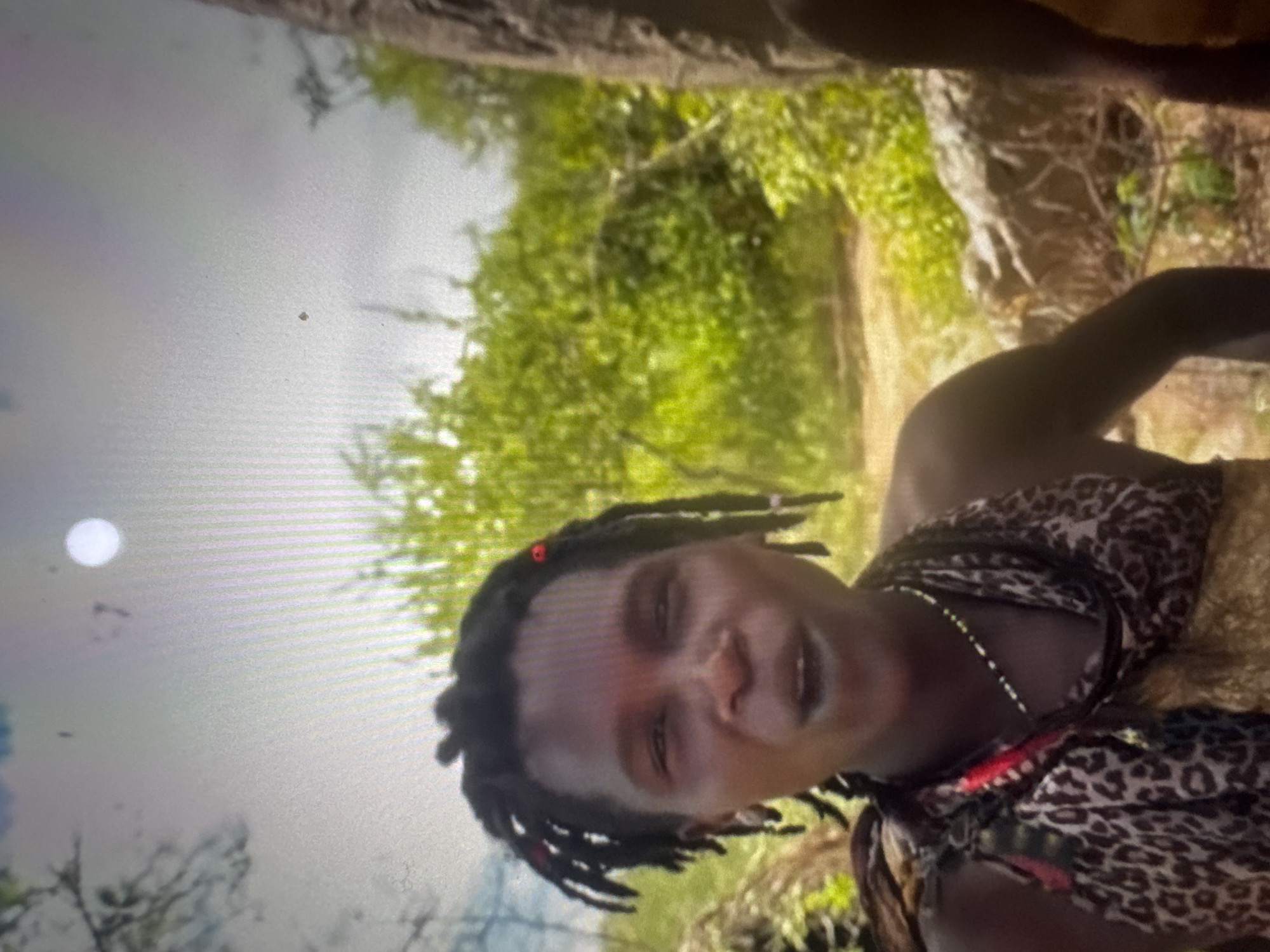 The image shows a medium shot of a person with dark skin and dreadlocks, wearing a leopard-print fabric around their shoulders and a beaded necklace. They appear to be speaking or reacting to something, with their mouth open and eyes looking slightly to the side. The background is lush green foliage and trees, suggesting an outdoor, natural environment. The lighting is bright, with a white circle visible in the upper portion of the frame, possibly a light source or lens flare. The image quality is somewhat grainy. The location is described as Unknown, Unknown.