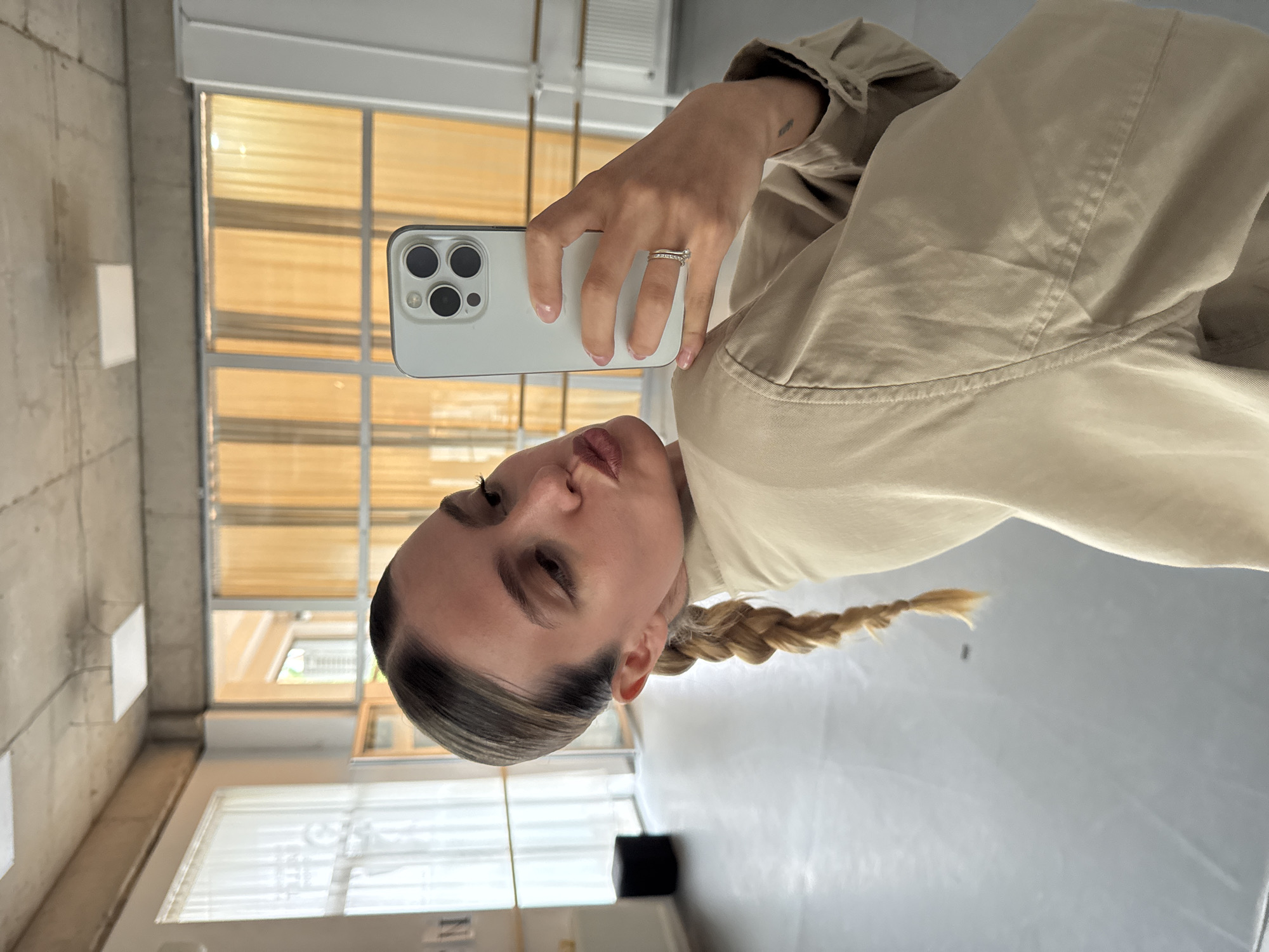 A woman with light skin and dark roots in her hair is taking a selfie with a white smartphone that has multiple cameras. She is wearing a light tan jacket and has her blonde hair braided. She is wearing a ring on her finger. The background consists of a room with windows and blinds. The location appears to be a room in Unknown, Unknown.