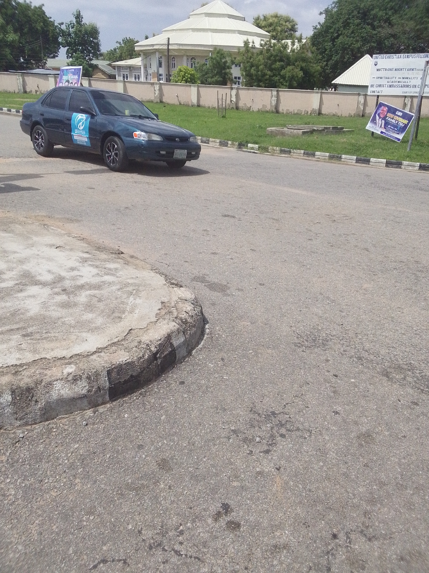 This media file depicts an outdoor scene in Jalingo, Nigeria, under what appears to be daytime with a cloudy sky. The primary focus is a dark-colored sedan car driving on an asphalt road. The car has decals on its side, one of which appears to be a blue fish logo with text "NISSAKAS" beneath it. Another visible decal on the car is a rectangular banner with text that includes "SOUND UP ALL URL".
In the background, a building with a distinctive white, multi-tiered roof structure is visible, surrounded by a concrete wall and trees. Adjacent to the building, on a patch of grass, are two signs. One sign is a large white banner with text including "UNITED CHRISTIAN CAMPUS FELLOWSHIP", "MOTTO ONE MIGHTY ARMY", and "BE CHRIST AMBASSADORS ON CP". Below this banner is a smaller, colorful sign featuring a portrait of a man and the text "COVIMANT FAMILY TSU". The road also features a concrete curb with black and white painted stripes. The overall impression is a daytime scene on a campus or institutional grounds.