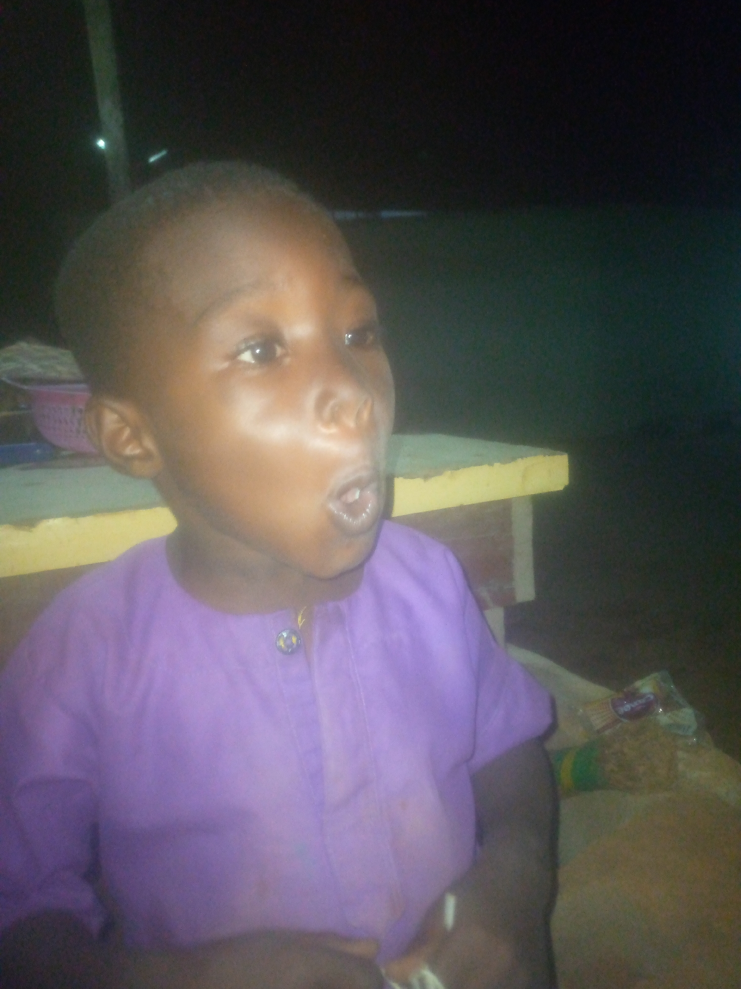 A medium shot depicts a young, dark-skinned child in Unknown, Unknown. The child, facing left, wears a purple button-down shirt. A light-colored surface is visible behind the child, possibly a table or bench. To the right of the child, there is a package of "Snacks" brand items. The background is dark and mostly obscured.