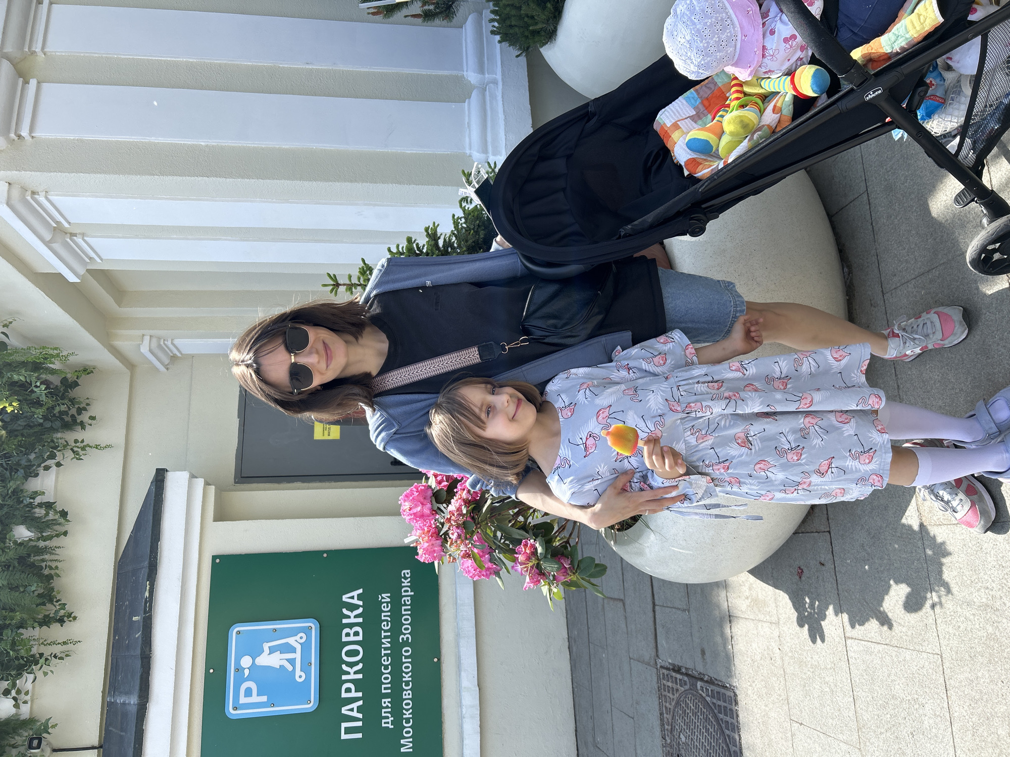 The image captures a bright, sunny outdoor scene featuring a woman, a young girl, and a baby in a stroller. The woman, wearing sunglasses, a black top, a blue jacket, and denim shorts, stands to the left, looking towards the camera with a gentle smile. Beside her, a young girl, also smiling towards the camera, is dressed in a light blue dress with a pink flamingo pattern, white tights, and light-colored sneakers. She holds a yellow-orange lollipop in her right hand. To the woman's right is a black baby stroller, a "Chicco" brand visible, containing a baby wearing a pink and white hat, a colorful blanket, and a striped soft toy.
The group is situated on a paved sidewalk, with a light-colored building featuring classic architectural details in the background. Lush green foliage and a large planter with vibrant pink flowers are visible to the left and center of the frame. The strong sunlight casts distinct shadows on the ground, indicating a clear day.
A prominent green sign on the left provides location cues. The visible Russian text reads "ПАРКОВКА" (Parking) at the top, and "для посетителей Московского Зоопарка" (for visitors of the Moscow Zoo) below. A blue square icon on the sign depicts a person on a scooter next to a "P" for parking.
The overall scene suggests a pleasant family outing, possibly at or near the Moscow Zoo, with the subjects posing happily for a photograph.