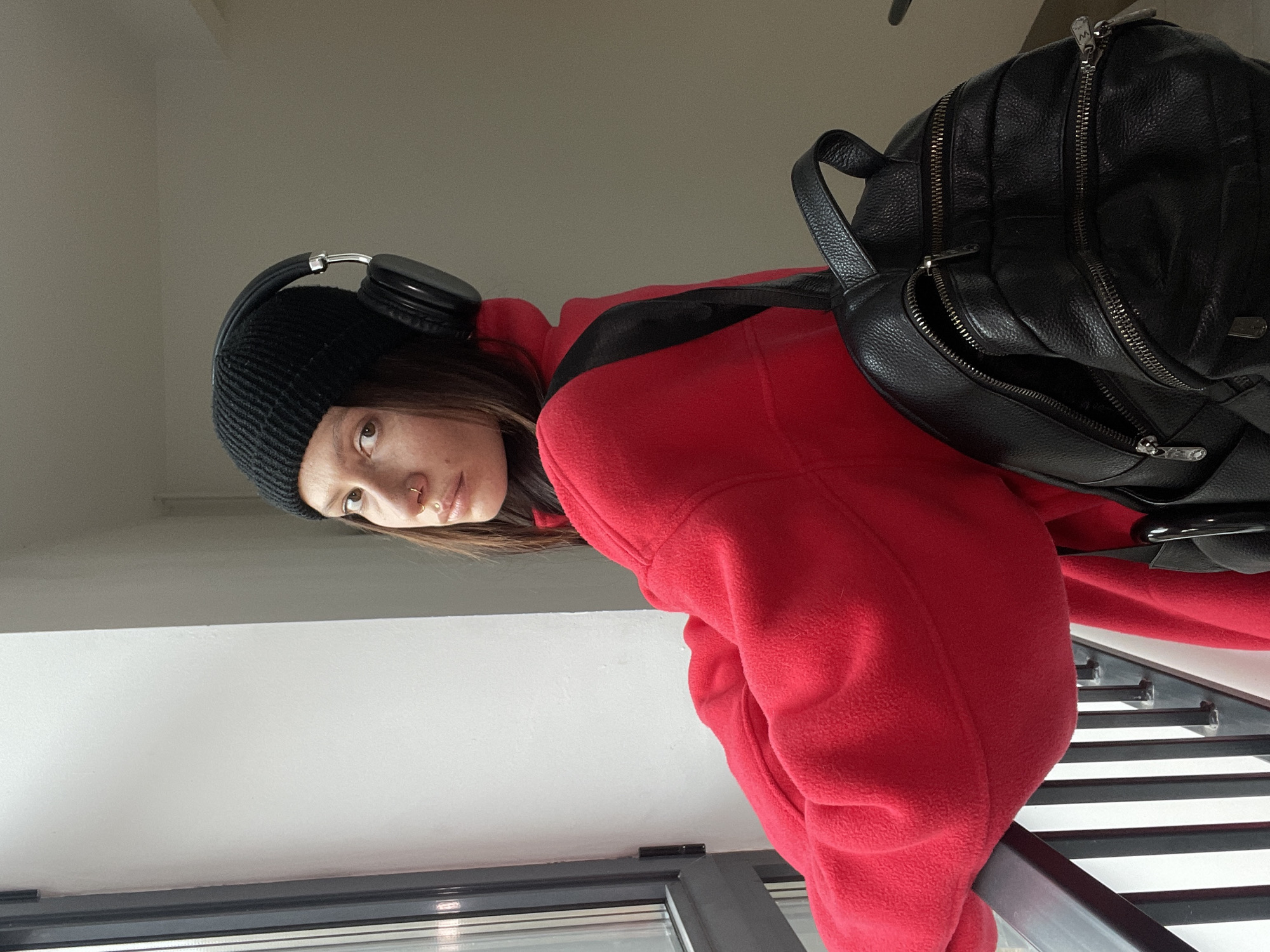 A woman wearing a black beanie, black headphones, and a red fleece jacket is carrying a black leather backpack. She is indoors, standing near a window with horizontal blinds. The exact location in Izhevsk, Russia cannot be determined without additional information.