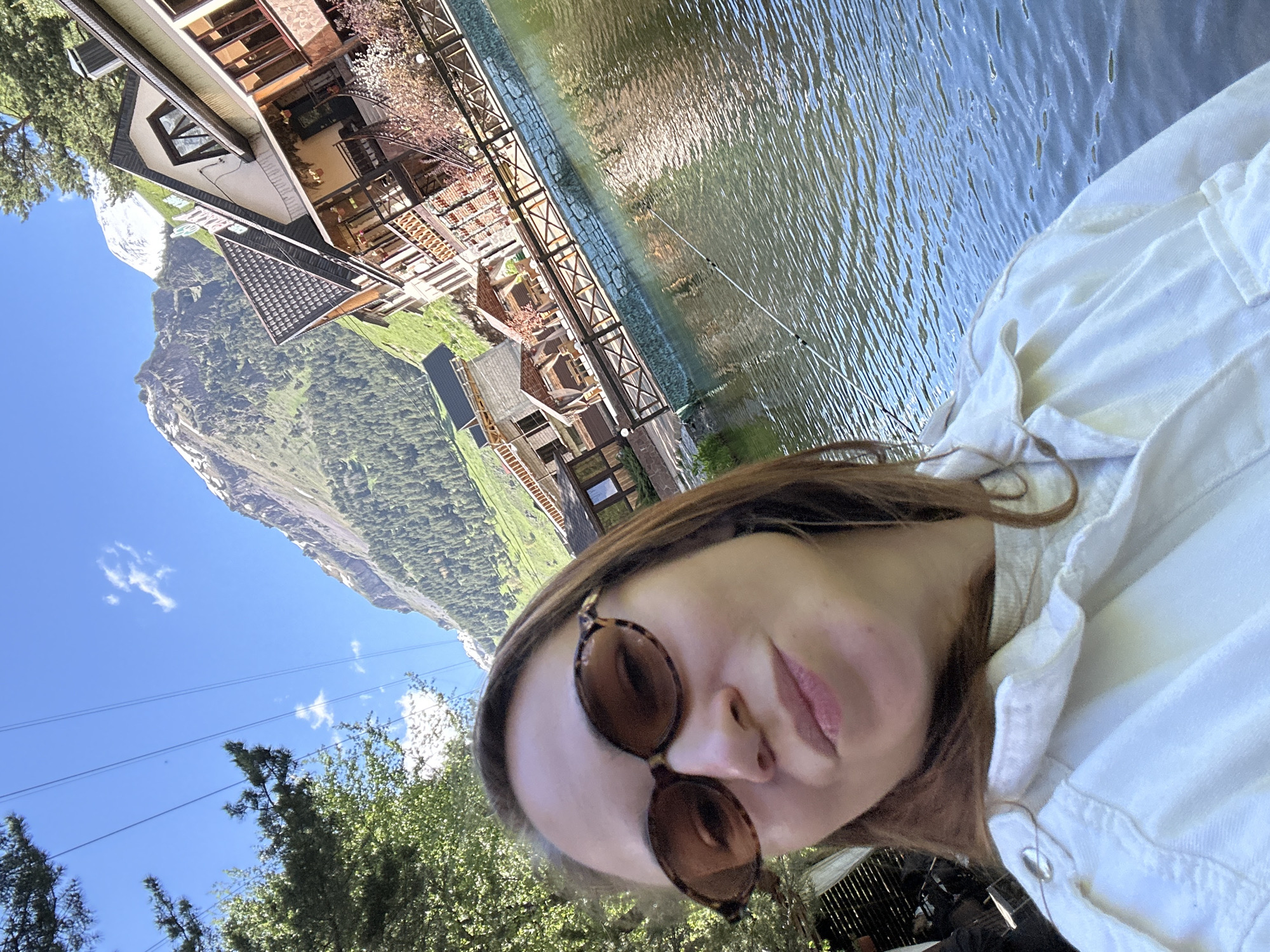 A woman is taking a selfie in front of a body of water with buildings and mountains in the background. The woman has brown hair and is wearing sunglasses and a white jacket. The water is calm and reflects the sky and surrounding scenery. Behind the water are several buildings with wooden accents, suggesting a resort or hotel. In the far background, a large, snow-capped mountain rises, characteristic of the Caucasus Mountains in the Elbrus region, near Terskol, Russia. The sky is blue with a few white clouds. There are trees on the left side of the image.