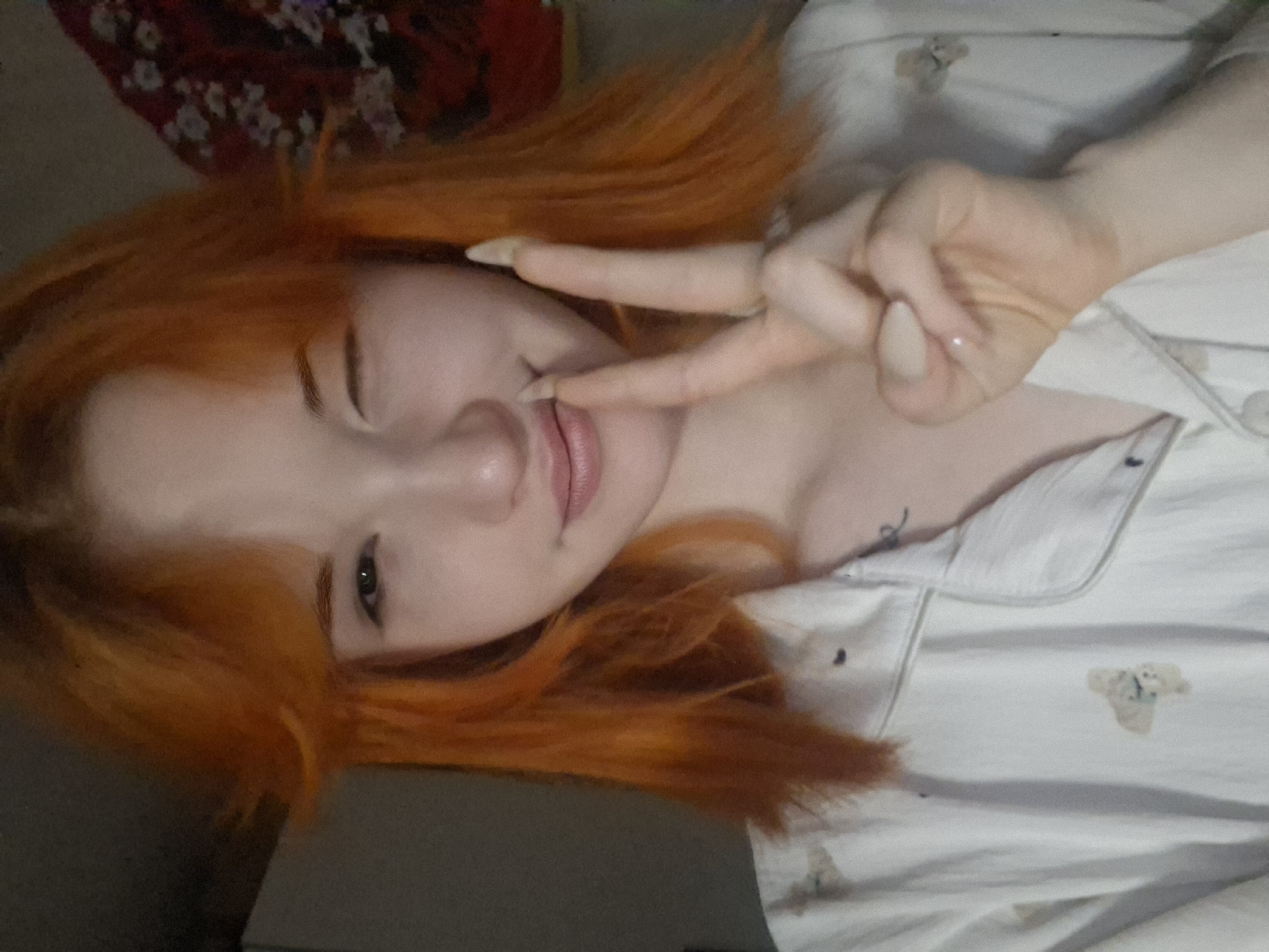 A young person with bright orange hair is depicted lying down, likely indoors in Chaikovsky, Russia. The subject is fair-skinned, with visible dark eyebrows and a dark iris in their open eye, while the other eye is playfully winking. They are making a peace sign with their left hand, with their manicured fingers touching their lips. Their nails are shaped and painted with a light, neutral polish. The person is wearing white pajamas that feature small, light-colored patterns, which appear to be teddy bears. In the upper left background, a dark fabric with a red and white floral pattern is visible, suggesting a bedspread or pillow. The image conveys a relaxed and playful mood. There is no visible text.