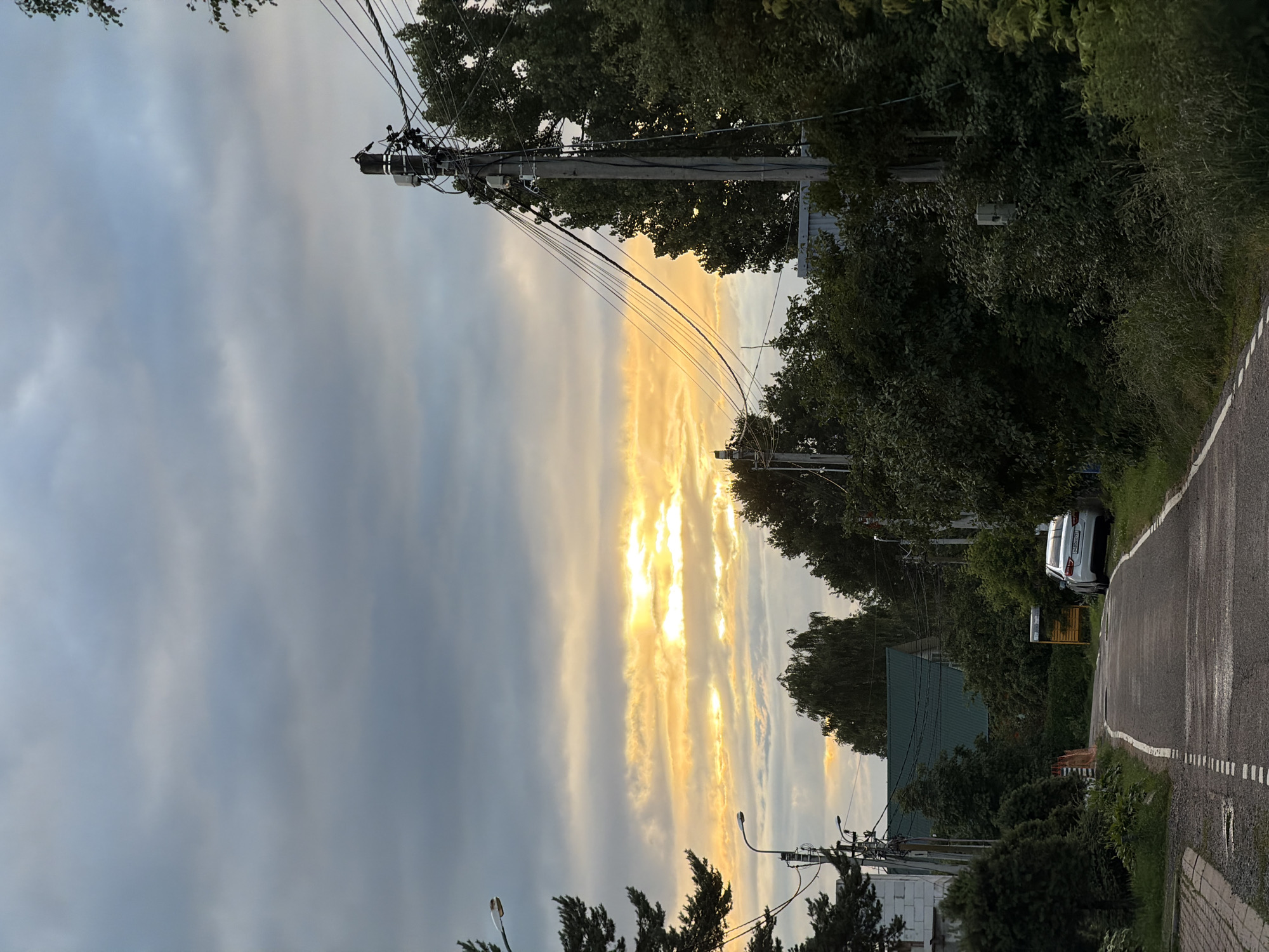 This media file depicts a rural roadside scene in Zimyonki, Russia, during what appears to be a cloudy sunset. The perspective is from ground level, looking down a paved road that curves to the right. Towering trees line the right side of the road, their dense foliage casting shadows. On the left side of the road, telephone poles with a complex network of wires are prominent.
The sky is filled with dramatic, wispy clouds that are illuminated by a bright, warm light, indicating the sun is setting. This creates streaks of yellow and orange through the grayer clouds. The overall mood is serene and somewhat overcast, with the sunset adding a touch of warmth.
Visible objects include a white car parked on the side of the road, several houses with green roofs, and streetlights. There are also what appear to be road signs and utility boxes attached to the telephone poles. The road itself has dashed white lines marking its edges and center.
The scene evokes a sense of peaceful, everyday life in a rural setting. The time of day is clearly dusk, with the light filtering through the clouds. The weather is cloudy but not actively raining. No people are visible in the frame. The location is hinted at by the provided context "Zimyonki (Zimenki, Zimenka), Russia." No discernible text is legible in the image.