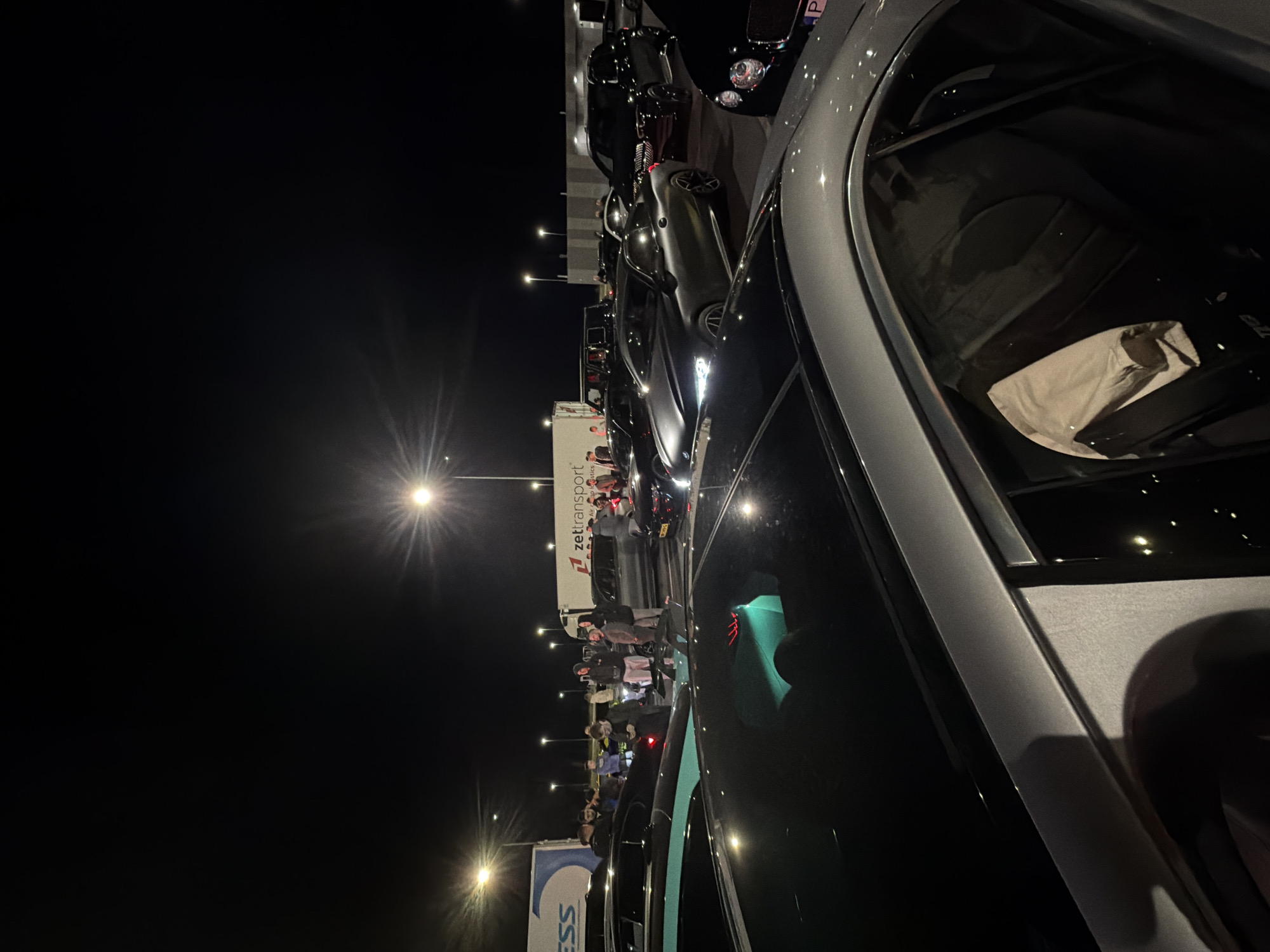 Here is a forensic description of the image:
The image, taken at night, depicts a gathering of people and cars in what appears to be a parking lot or rest area. The scene is lit by overhead streetlights, casting a bright glow with some glare. A truck with the logo "zet!transport" is visible in the background, around which a group of people seems to be gathered. Several cars, primarily dark-colored, are parked closely together, suggesting a car meet or similar event. The angle of the shot is close-up, capturing the side and windshield of a silver/gray car, likely where the photograph was taken from. Reflections are visible on the car's windows. No specific city can be determined from the visual information provided.