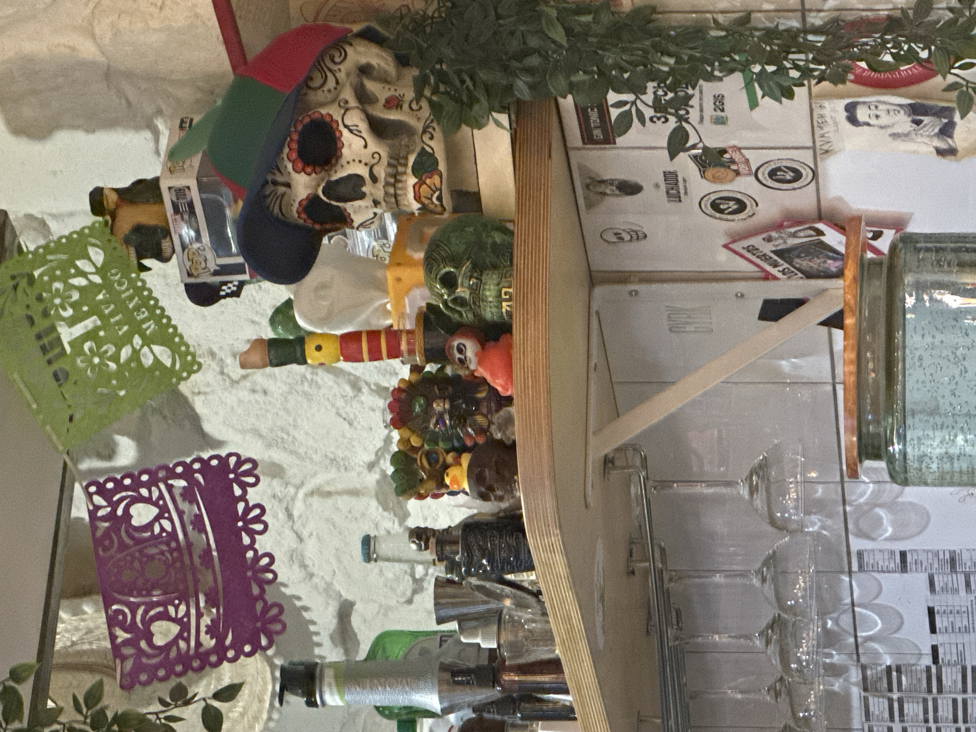 This image, presented sideways, depicts a densely decorated shelf or bar area with a strong Mexican theme, set against a textured white wall.
The main focal point is a large, ornate sugar skull wearing a colorful cap, positioned on a light-colored wooden shelf. Nearby, a smaller, dark green decorative skull and various colorful figurines and objects are arranged, including what appears to be a small "FUNKO" pop toy in a box. Artificial green foliage hangs over these items.
Two pieces of *papel picado* (cut paper decorations) are visible. One, green, displays the text "VIVA MEXICO" and "15". The other, purple, features intricate heart and floral designs.
Beneath the main shelf, several clear, stemmed glasses (resembling margarita or martini glasses) hang upside down on a metal rack, casting distinct circular shadows. A variety of bottles, likely containing beverages, are also present on the shelf, along with a large glass jar or dispenser.
In the background, behind the main display, stacked cardboard boxes are visible. Identifiable text on these boxes includes "2GIS", "LUCHADORE", and "CORK". A small paper cutout or picture of a person's profile is taped to one of the boxes.
No people are present in the scene. The decorations, particularly the sugar skulls and "VIVA MEXICO" text, strongly suggest a celebration or establishment with a Mexican theme, possibly related to Mexican Independence Day or Day of the Dead, located in Novosibirsk, Russia.