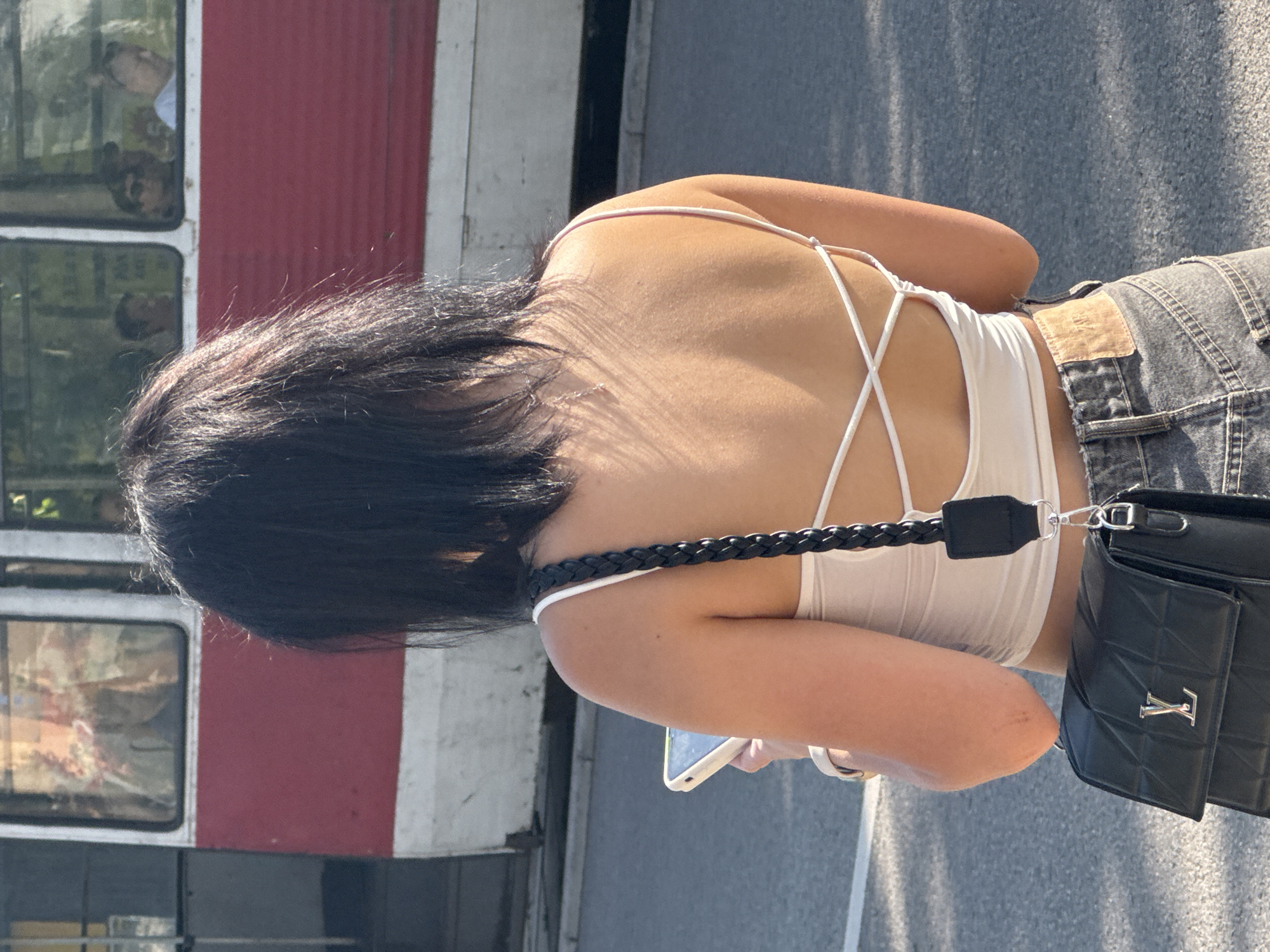 A young woman with dark hair is seen from behind, standing on a paved street, possibly in the city of Kryvyi Rih, Ukraine. She is wearing a white backless top with criss-cross straps and denim jeans. A black quilted handbag with a silver logo is slung over her shoulder, and she is holding a white smartphone in her left hand. In the background, a red and white tram with visible passengers inside is stationary. The lighting suggests it is daytime, with sunlight casting shadows on the ground.