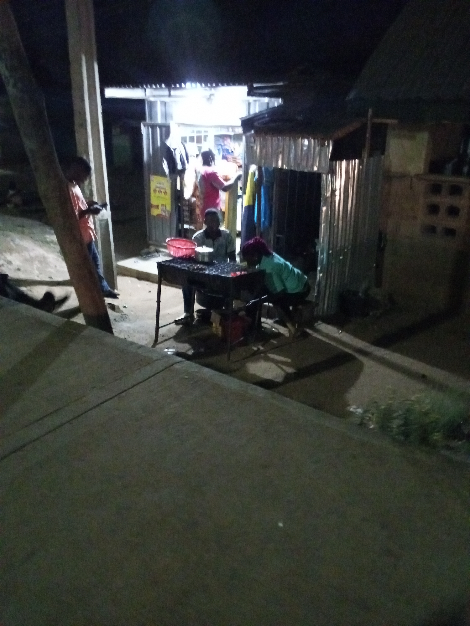 Here's a detailed description of the image:
**Scene:** The image shows an outdoor nighttime scene in what appears to be a densely populated, urban area.
**People:** There are multiple people in the image.
* A person is standing next to a tree and a wooden pillar, looking at a device that they are holding.
* A person is seated at a metal grill, presumably cooking food. A red plastic bowl is on the grill as well as a metal pot with a lid.
* Another person in a turquoise shirt and a reddish head covering is bent over next to the grill.
* Two more people can be seen inside the shop.
**Objects:**
* A metal grill stand with items being cooked.
* Two adjacent small shops/structures. One appears to be a permanent shop with corrugated metal sides, a door, and a bright light emanating from inside. A sign is on the side. The other is constructed of corrugated metal and appears to be a food preparation area.
* A tree and wooden pillar that frame the scene on the left side.
* A dark paved road or sidewalk dominates the foreground.
**Actions/Interactions:**
* Cooking is underway at the grill.
* A person is standing next to a tree, looking at something that is on their phone.
* The scene suggests a busy, possibly late-night food vending operation.
**Environment:**
* The lighting is dim but partially illuminated by the artificial lights of the shop.
* The area appears to be a roadside location with a concrete sidewalk or road.
* The overall setting implies a commercial or residential neighborhood.