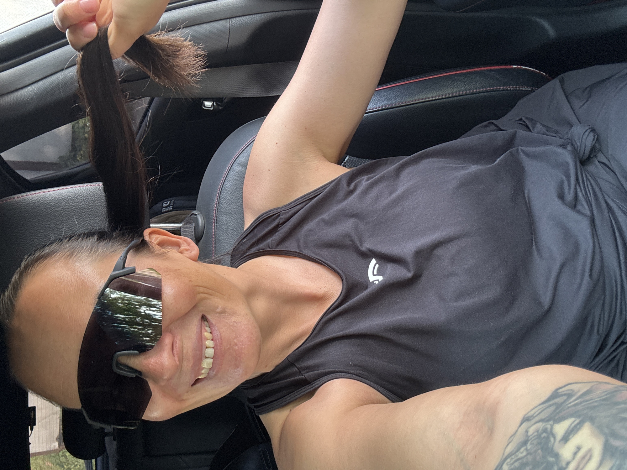 A selfie depicts a woman sitting in a vehicle, possibly a car. She is fair-skinned and has dark hair pulled back, wearing sunglasses and a black sleeveless athletic shirt. Her left arm is raised, holding a clump of cut dark hair in her hand. The woman is smiling. The interior of the car features black leather seats with red stitching. The dashboard is visible. The photo appears to be taken outdoors during the daytime, with trees and foliage reflected in the sunglasses. There is no information available in the image to indicate its location in the city of Chaikovsky, Russia.