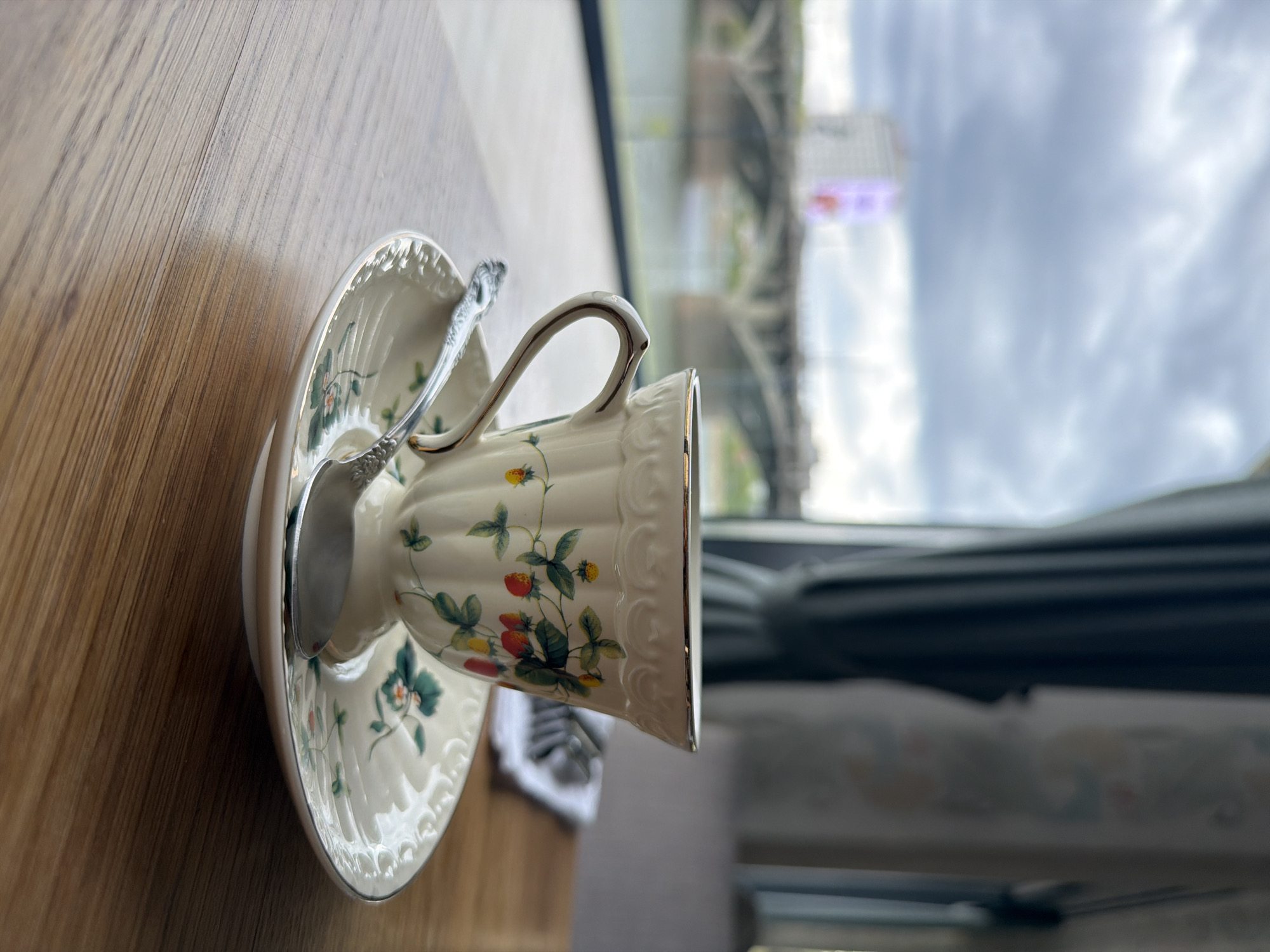 A high-angle, tilted, close-up shot shows a teacup and saucer, with a spoon resting on the saucer, on a wooden surface. The cup and saucer are decorated with a strawberry pattern and gold trim. The background reveals a blurred view of an indoor space with a window. Outside the window, structural elements are visible, appearing to be parts of a bridge or elevated structure. Given the location information (Krasnoyarsk, Russia), the structure seen through the window is likely the Vinogradovsky Bridge spanning the Yenisey River.