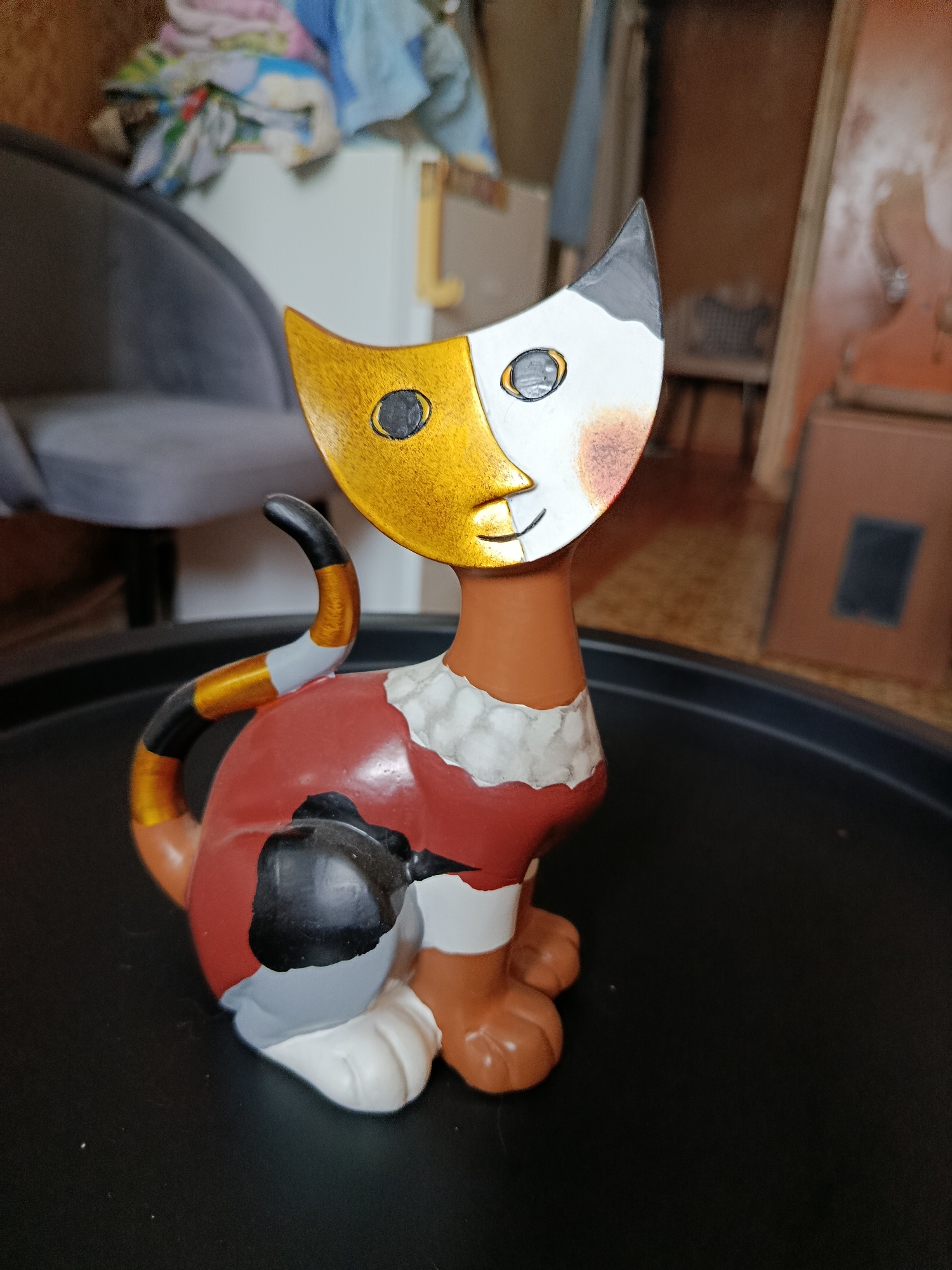 A decorative ceramic cat figurine is displayed on a black, circular tray. The figurine is anthropomorphic with a stylized face featuring half gold and half white with black and grey accents. It has a brown neck, a red body with black and white patches, and grey paws with white tips. The tail is curved and striped with gold, black, and white. The figurine appears to be a home decor item. The background is blurred but suggests an indoor environment with furniture, including a grey chair, and possibly a dresser or cabinet. The overall impression is of a still life arrangement within a domestic setting. The location of the image in the city of Vladimir, Russia is not discernible from the visual information provided.