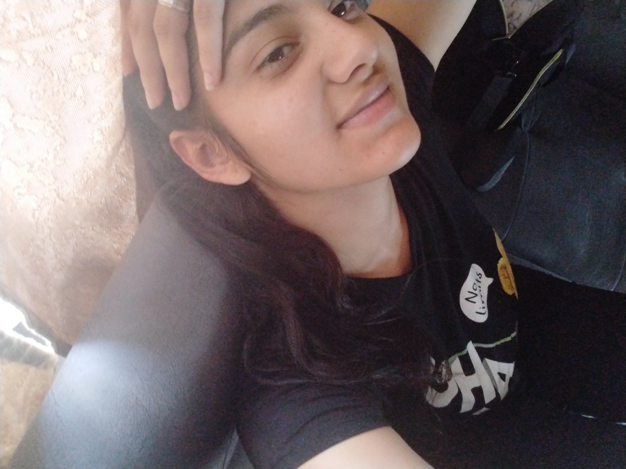 A close-up, over-the-shoulder selfie shows a young South Asian woman with long, dark hair wearing a black t-shirt with white text on it. She is resting her head on her left hand, which is propped on a textured, light-colored surface. Her right hand is visible near her ear. She is looking upwards and to her left. The background is dark, with a hint of a black bag visible on the right side. The scene appears to be indoors, possibly a living room or bedroom.