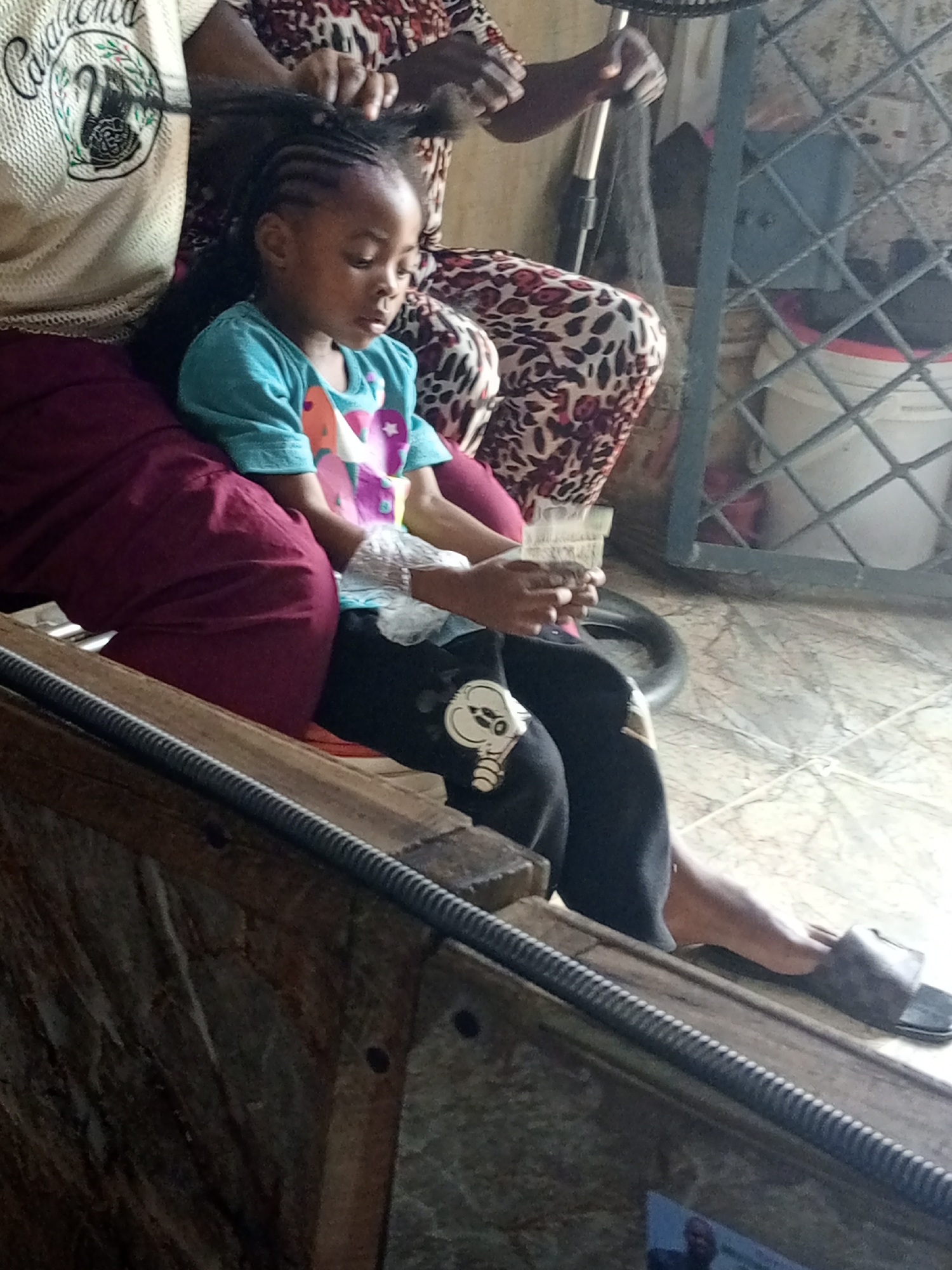 A young Black girl, appearing to be about five years old, sits on a maroon cushion. She is having her hair braided by an unseen person. The girl is wearing a turquoise t-shirt with colorful graphics and black pants with a white cartoon character on the knee. Her hands are clasped together. To the right of the girl, a gray fan is visible. The background consists of a tiled floor and a metal grate. A pair of dark brown slippers rests on the floor to the girl's right.