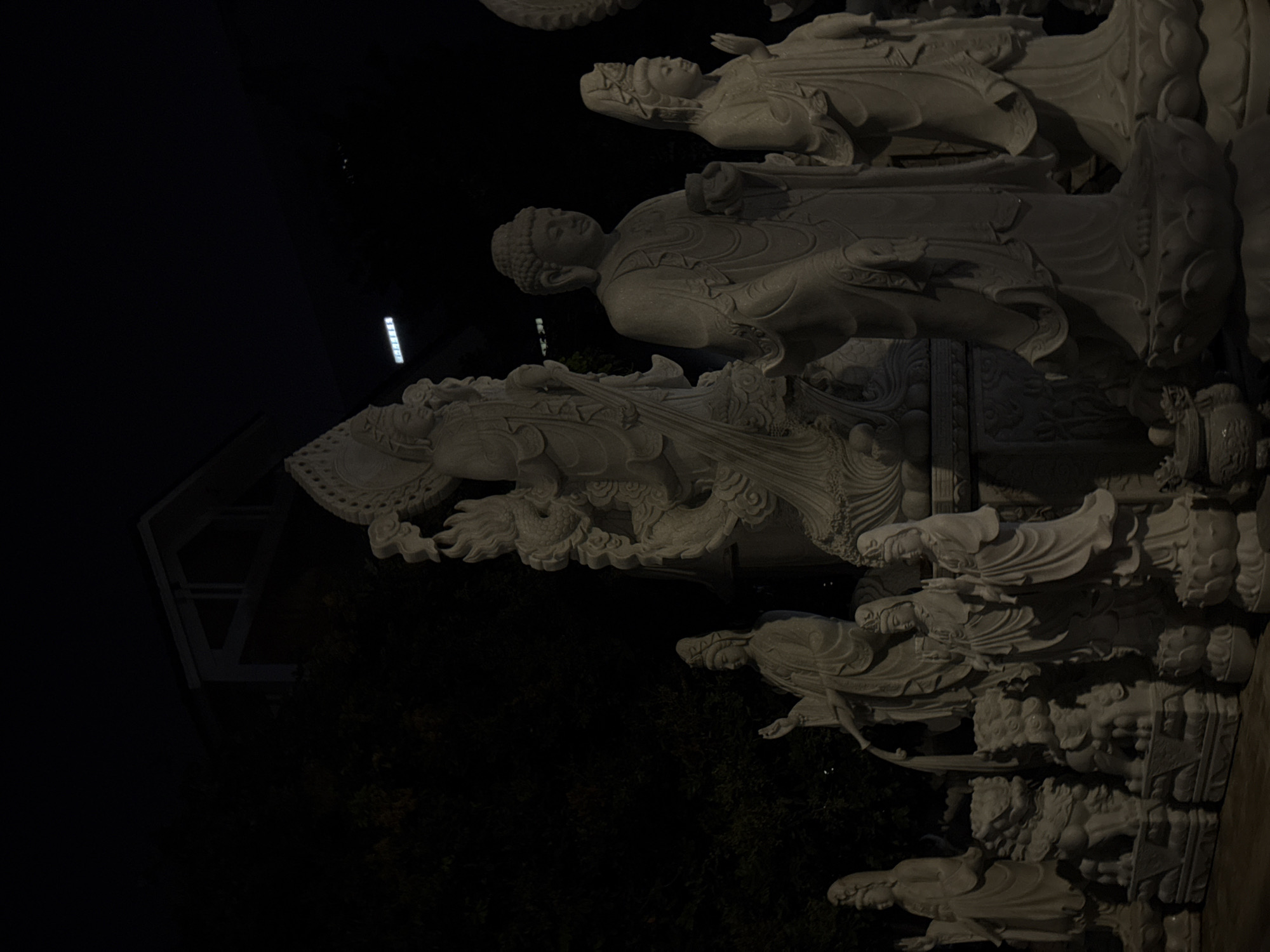 The image captures a series of white marble statues depicting figures, likely Buddhist deities, at night in Đà Nẵng, Vietnam. The statues are arranged vertically. A dragon statue is most prominent. Other statues appear to represent human-like figures, possibly Buddhas or Bodhisattvas. The background is dark, with vague outlines of trees and a building visible. A rectangular light source illuminates part of the statue arrangement. The overall scene is dimly lit and focused on the intricate carvings of the statues.