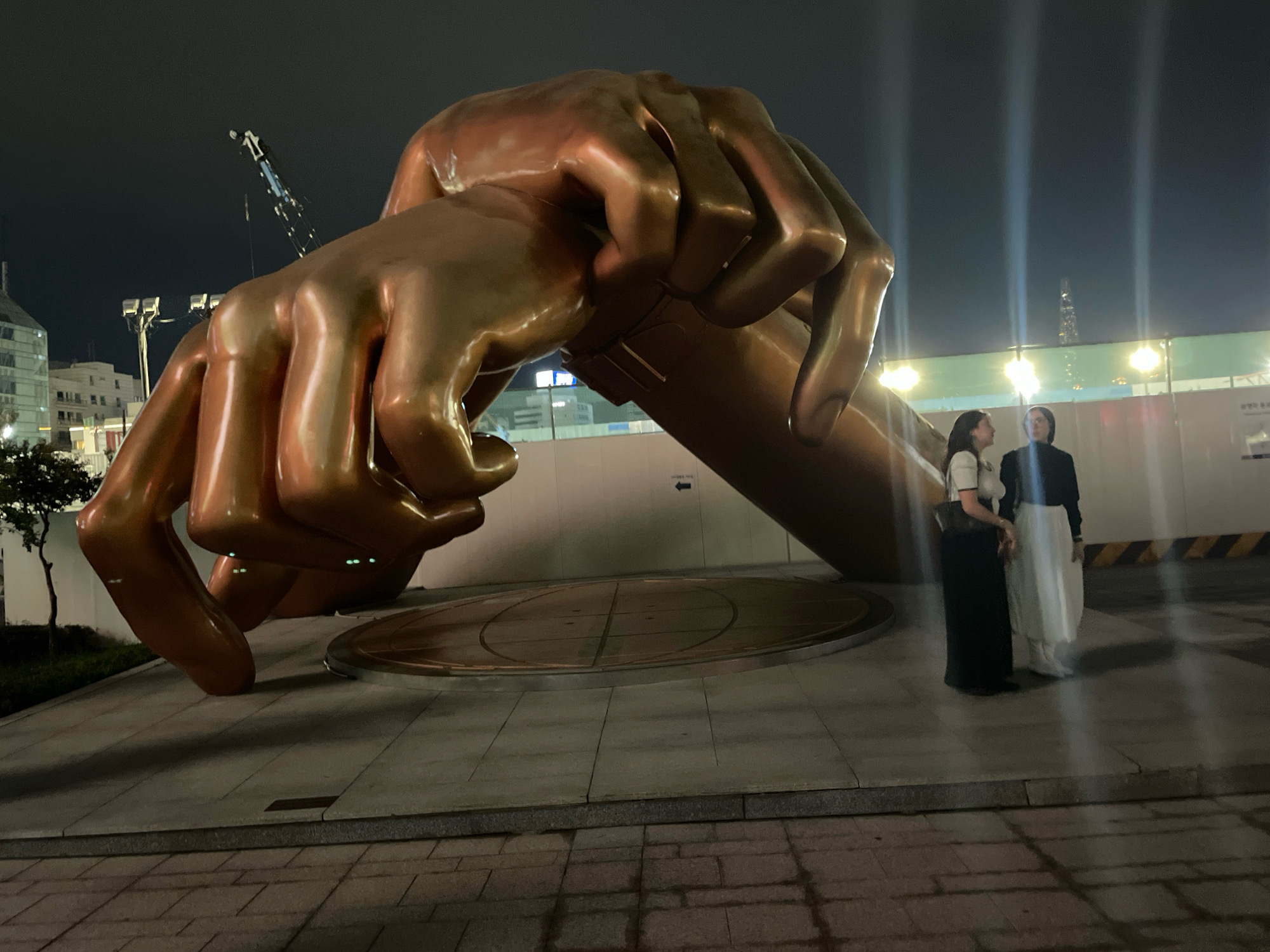 At night in Seoul, South Korea, a large bronze sculpture of two clasped hands dominates the scene. The sculpture is positioned outdoors, with a dark sky overhead. Two individuals stand near the base of the sculpture, appearing to observe it. One person is wearing a white top and a long black skirt, while the other is dressed in a black top and a long white skirt. The ground around the sculpture is paved with concrete tiles. In the background, buildings and construction fencing are visible, along with lighting that illuminates the area. A crane is also visible in the far background, suggesting ongoing construction or development in the vicinity.