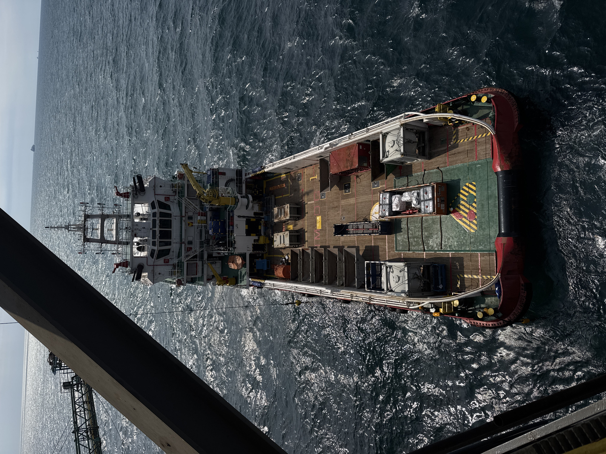 An aerial, high-angle shot captures a large, modern supply vessel with a red and white hull floating on a dark, choppy sea under a bright, hazy sky. The vessel is laden with various cargo, including several grey containers, orange crates filled with bagged goods, and stacks of what appear to be wooden pallets. The deck is a mixture of wood and green painted areas with yellow and black hazard stripes. A large, dark metal beam juts into the frame from the left, partially obscuring the view of the vessel and a metal walkway extending from an unseen structure. In the distance, several other ships are visible on the horizon, suggesting the vessel is in a busy shipping lane or offshore industrial area. The water reflects the sunlight, creating a shimmering effect. The scene is devoid of any visible human presence.