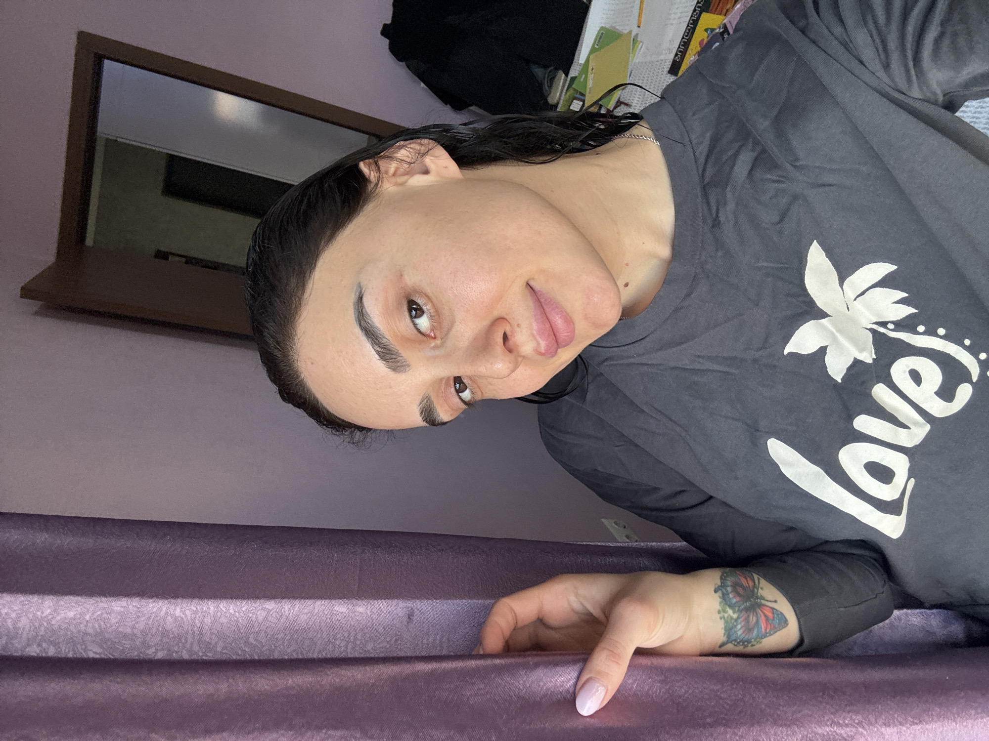 A woman with dark, damp hair and dark eyebrows is captured in a close-up, sideways shot, lying on a purple, textured surface, possibly a bed or couch. She is looking towards the camera with a subtle, slight smile or smirk. Her skin appears natural with no visible makeup.
She is wearing a dark grey long-sleeved shirt that features "Love" written in white cursive text, alongside a white floral or leaf design on the front. On her left forearm, a prominent, colorful butterfly tattoo is visible, displaying shades of red, blue, and green. Her fingernails have a light pink or ombre polish.
The background shows a light purple wall. To her upper left, a dark-framed mirror or window reflects a dim, indistinct area. Further back, on a shelf or surface, some papers or documents are scattered. The overall setting appears to be indoors, likely a bedroom or living area. The lighting suggests ambient indoor illumination. While "Tbilisi, Georgia" is provided as context, there are no specific visual cues in the image itself to confirm this location.