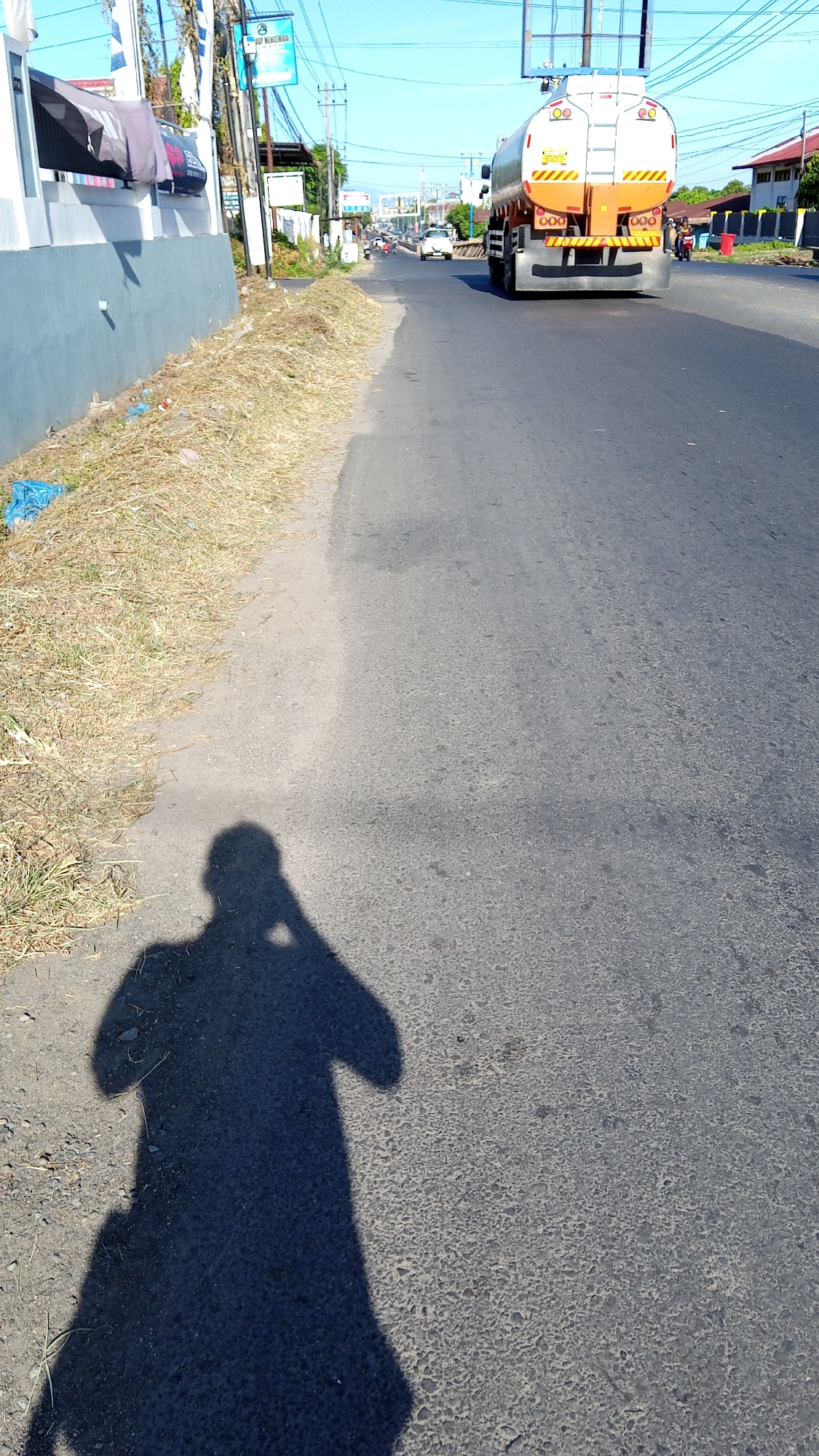 In the city of Bireuen, Indonesia, on a clear, sunny day, a scene unfolds along a paved road. The road, appearing slightly aged and textured, runs towards the background, where other vehicles and buildings are visible. To the left, a low, grey concrete wall lines the edge of the road, with dry grass and some scattered plastic debris at its base. Beyond the wall, a building with a white facade is partially visible. On the road, a large orange and white tanker truck is traveling away from the viewer, occupying the right lane. A white car is in front of it, and other vehicles and motorcycles are further down the road. The shadow of a person holding a phone is cast prominently on the road in the foreground, indicating the photographer's presence. The sky is a bright, cloudless blue.