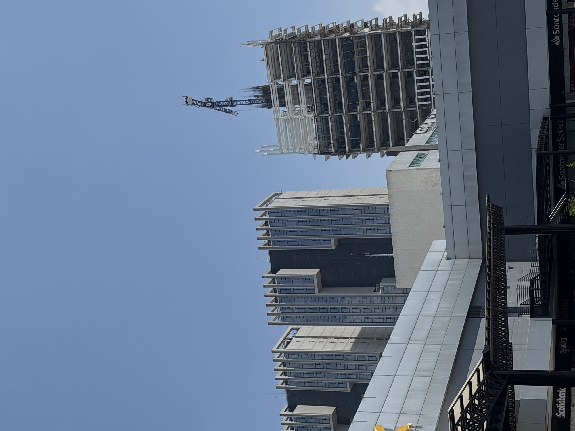 The image captures a view of buildings in San Bernardino Tlaxcalancingo, Mexico. A partially constructed high-rise building with a crane is visible. A completed building with a multi-level facade stands adjacent to it. Other buildings are also visible. Street-level elements, including signage for "Scotiabank" and "Santander Select," are partially visible on the right edge of the frame. The sky is clear and blue.