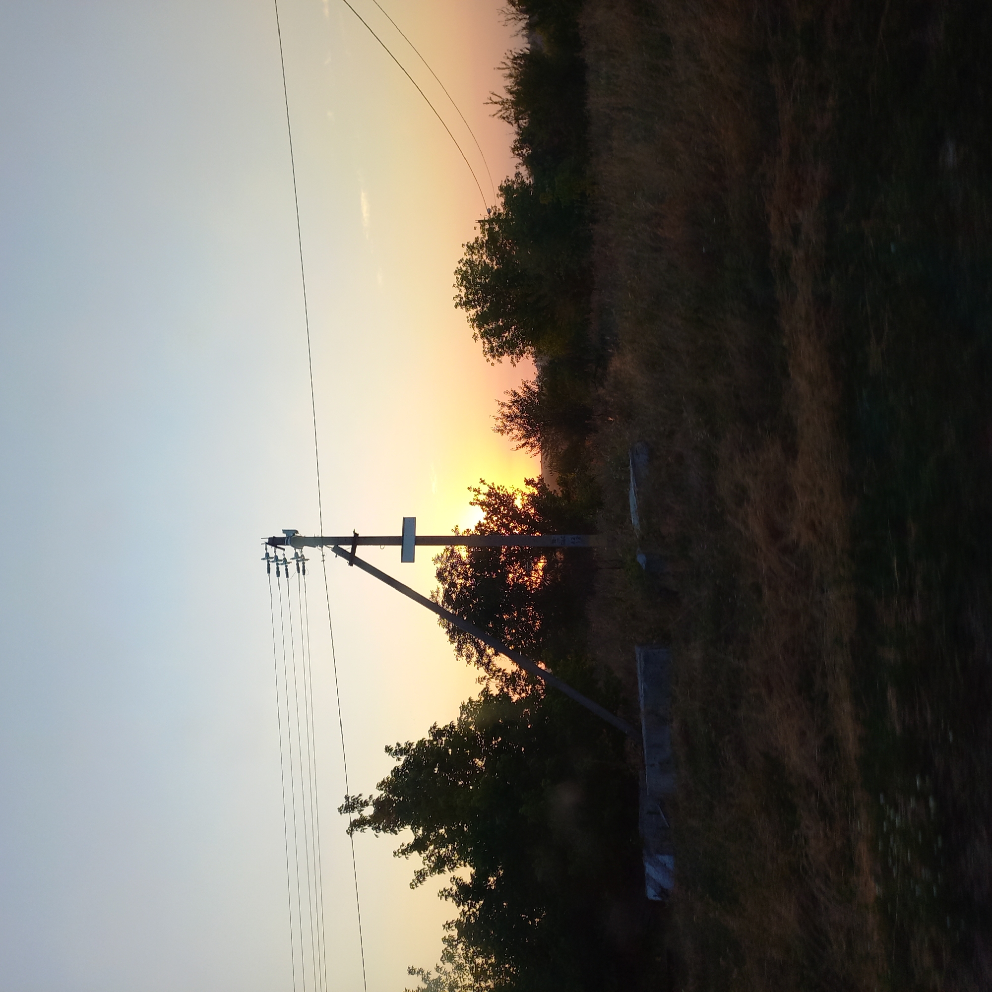 The image depicts a rural scene at dusk in the city of Soniachne, Ukraine. A wooden utility pole stands prominently in the center, supporting electrical wires that stretch across the frame. The setting sun is visible behind a cluster of trees, casting a warm, orange glow across the sky and illuminating the silhouette of the foliage. The foreground consists of dry, tall grass and what appears to be a concrete foundation or structure, suggesting a rural or undeveloped area. The overall atmosphere is tranquil and serene, characteristic of a sunset in the Ukrainian countryside.